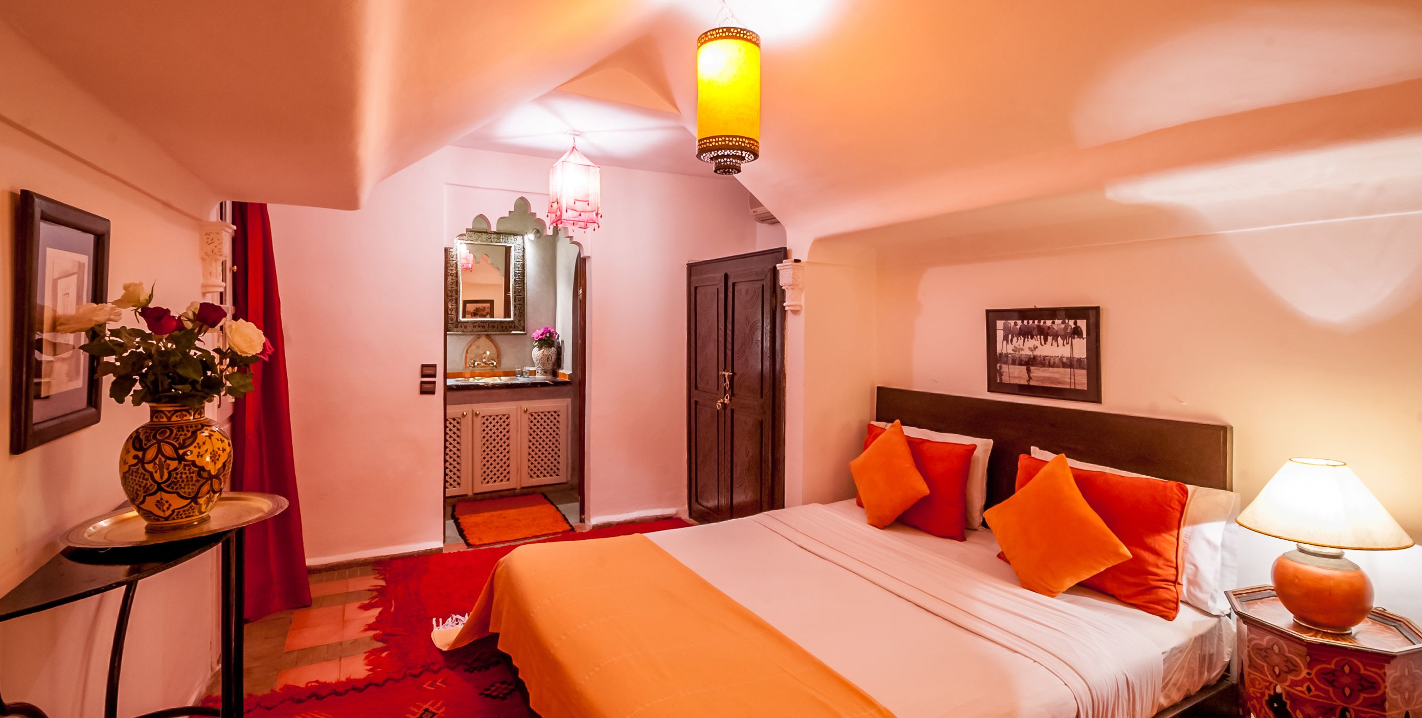 A hotel room with a bed, a nightstand, a lamp, a vase with flowers, a mirror, a door, and a small table.