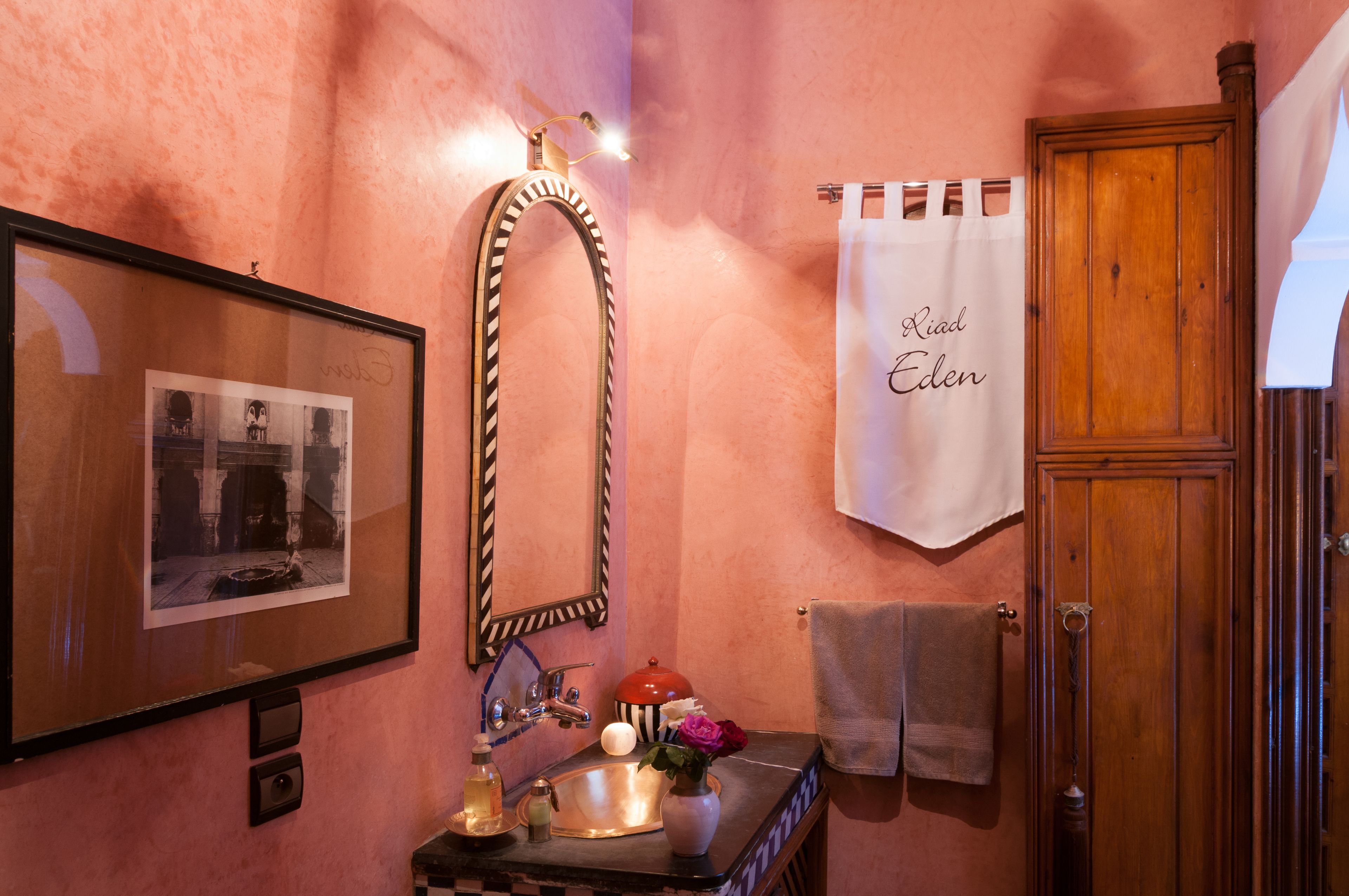 Une salle de bain avec une photographie encadrée, un miroir, un lavabo agrémenté d’un vase de fleurs et une serviette accrochée au mur.