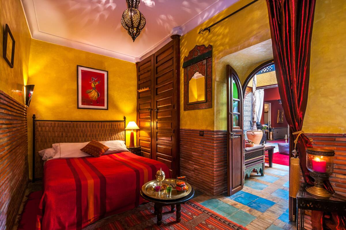 Una habitación con cama, una mesita con bandeja, un espejo y vista a otra habitación a través de un arco.