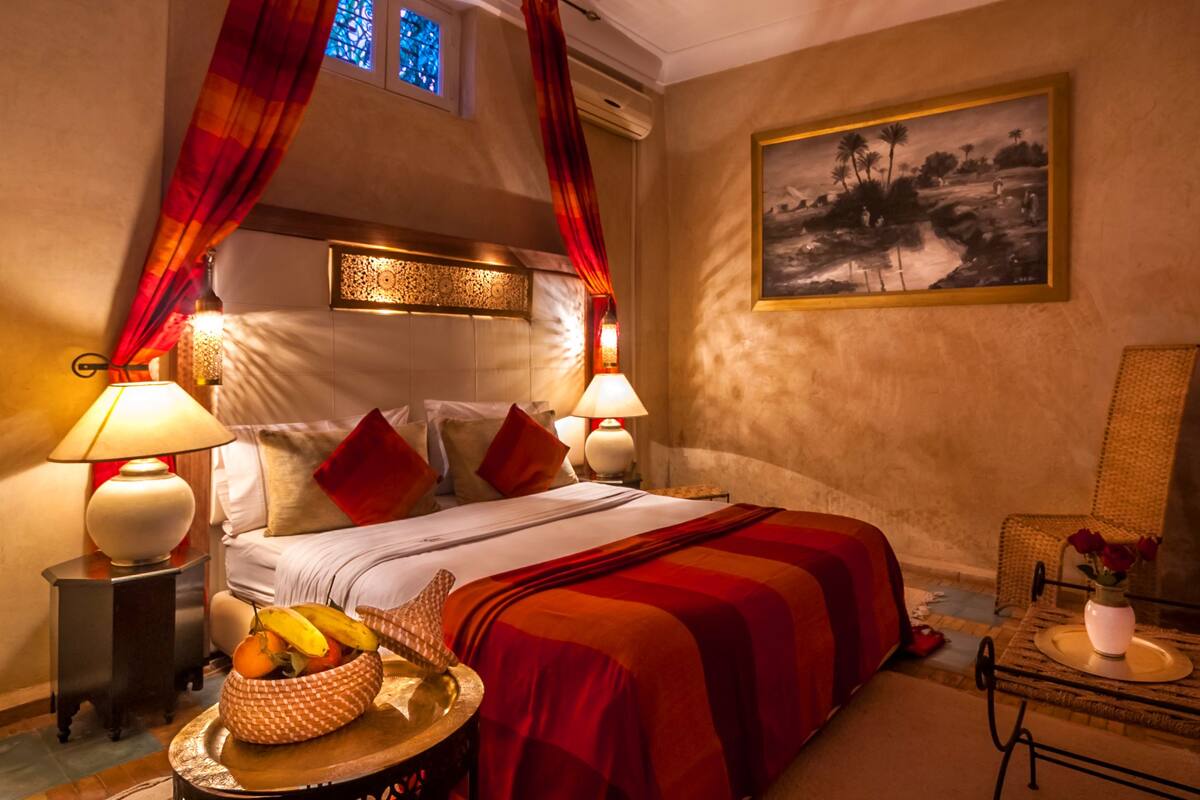 Habitación de hotel con cama, dos lámparas de noche, un cuadro en la pared, una mesita con un jarrón y flores, y una ventana con cortinas.