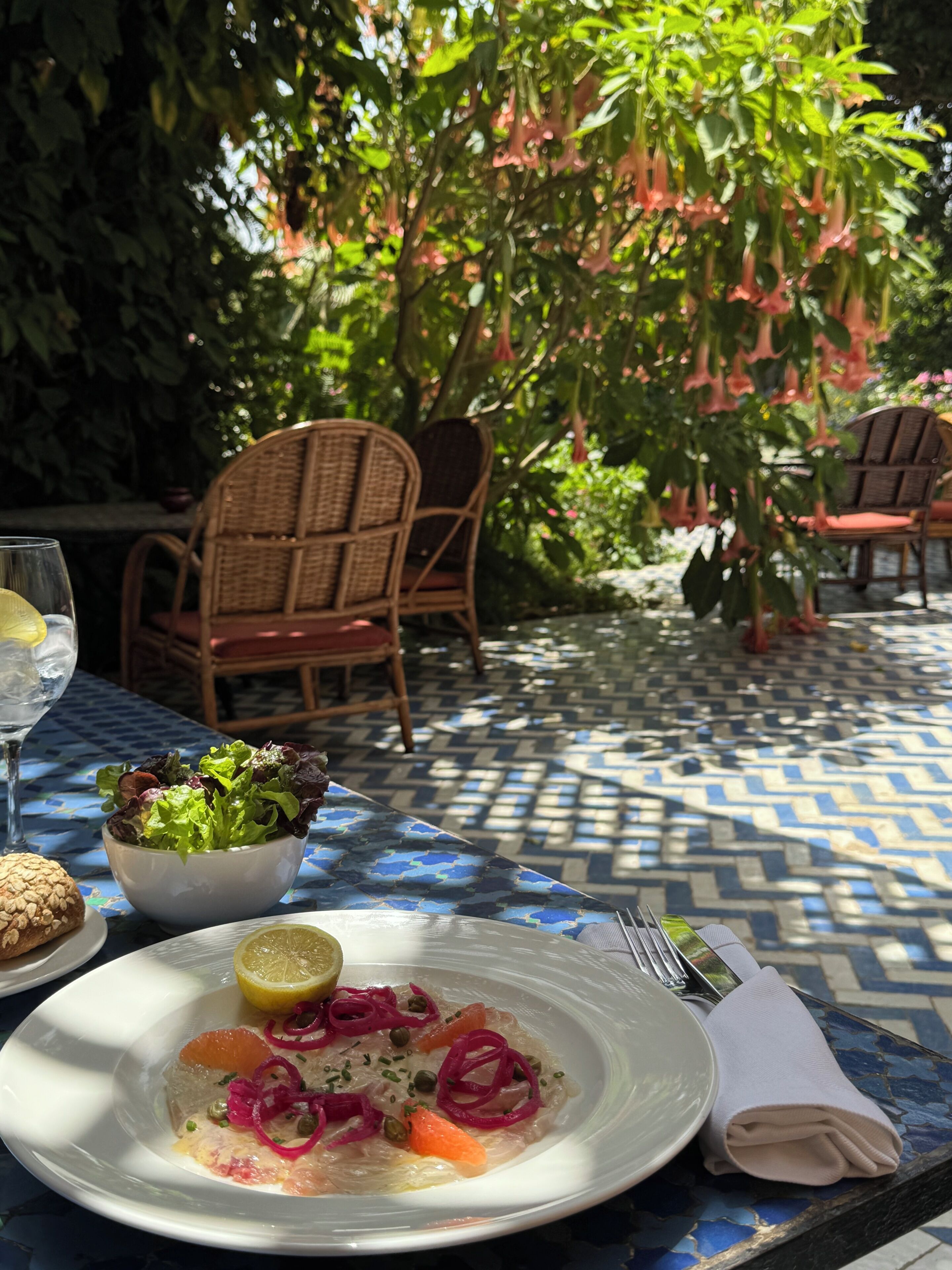 Ein Tisch mit einer blau gemusterten Tischdecke, einem Teller mit Essen und einem Glas Wasser, dazu Korbstühle.