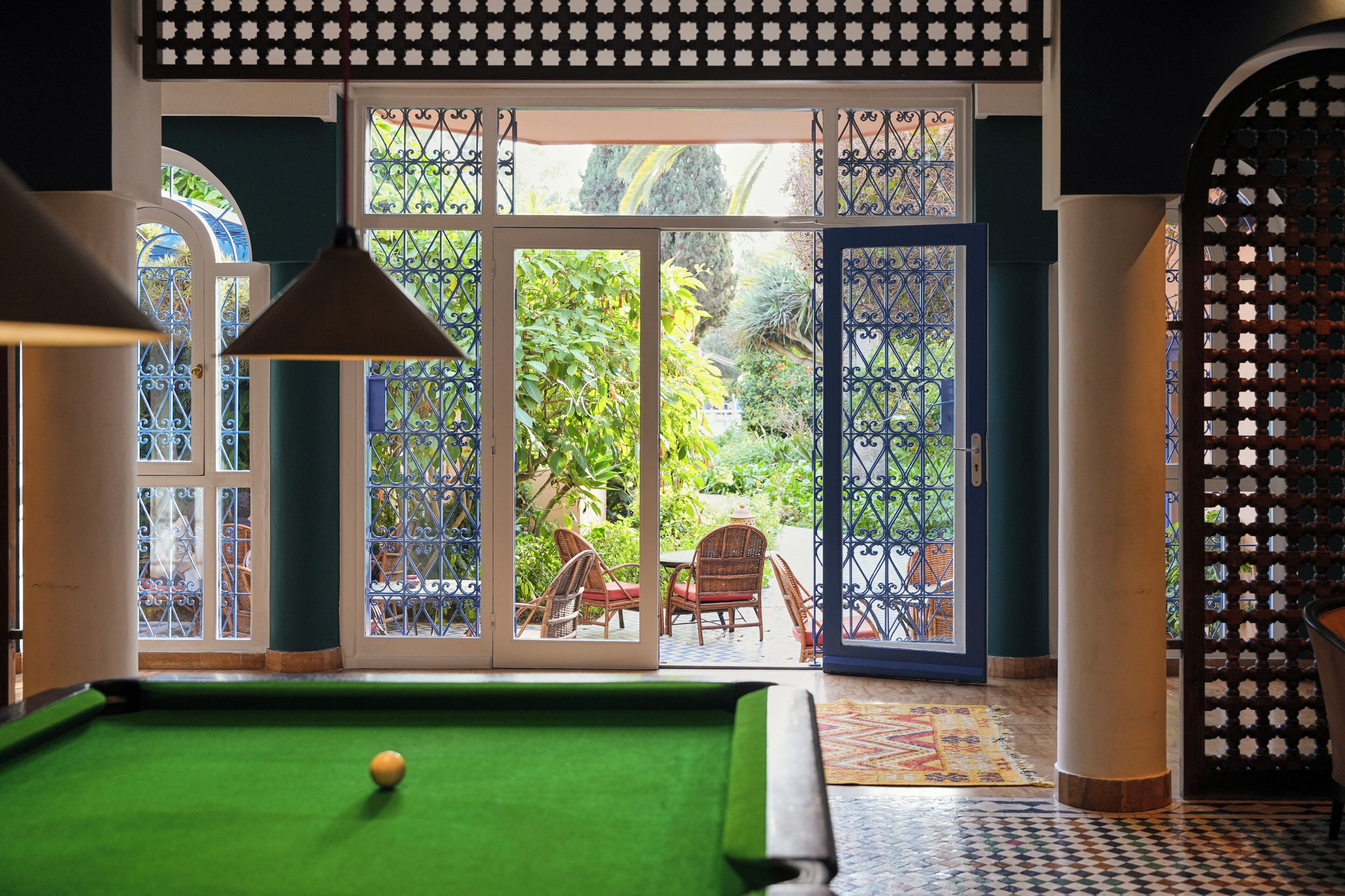 A pool table with a green felt surface and a single yellow ball, surrounded by a patterned floor, a door with glass panels, and a view of an outdoor patio area with chairs and plants.