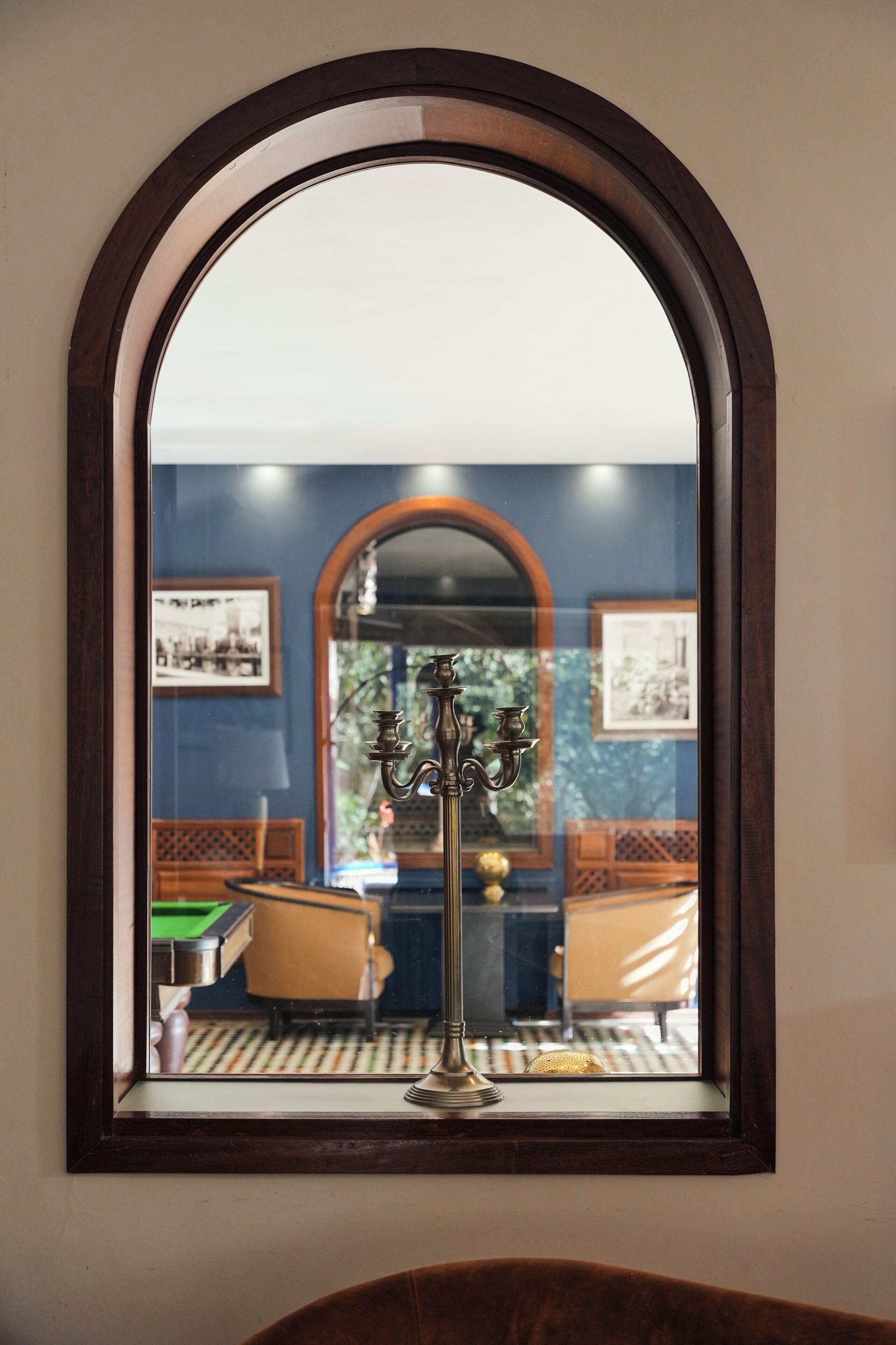 A mirror with a dark frame reflecting a room with a green table, a chair, and a candelabra.