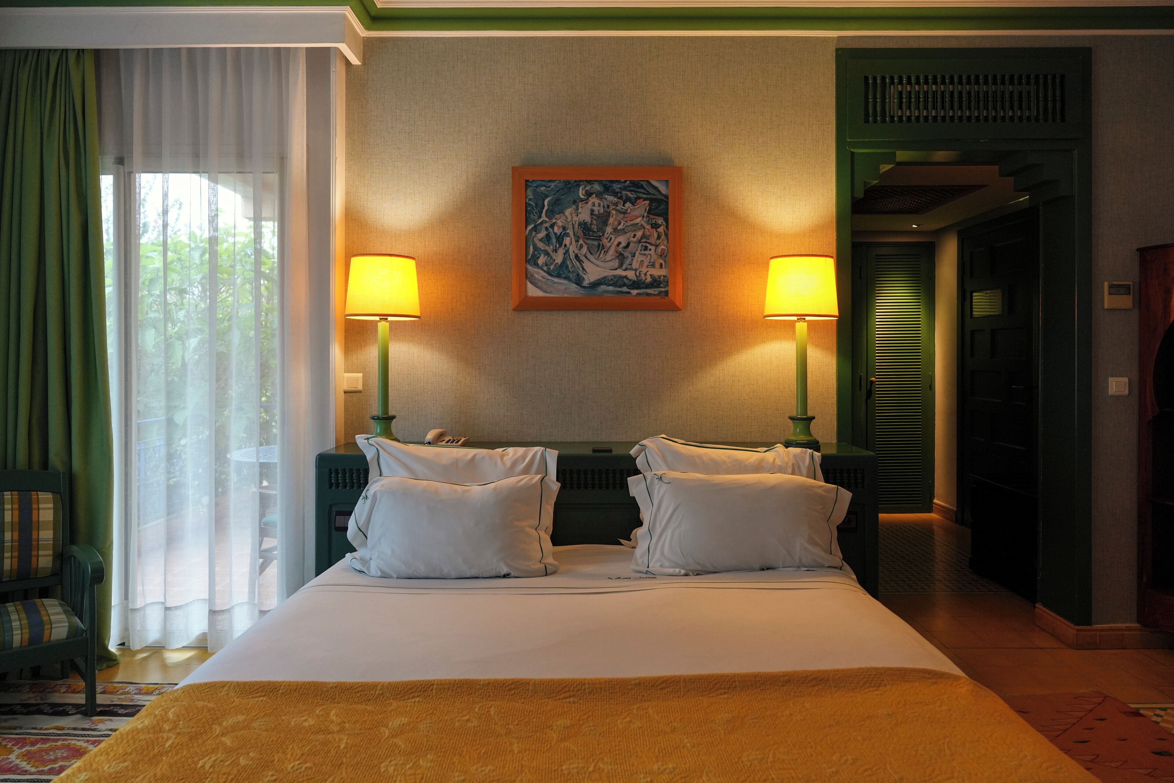 A hotel room with a bed, two bedside lamps, a painting on the wall, and a view through a window with curtains.
