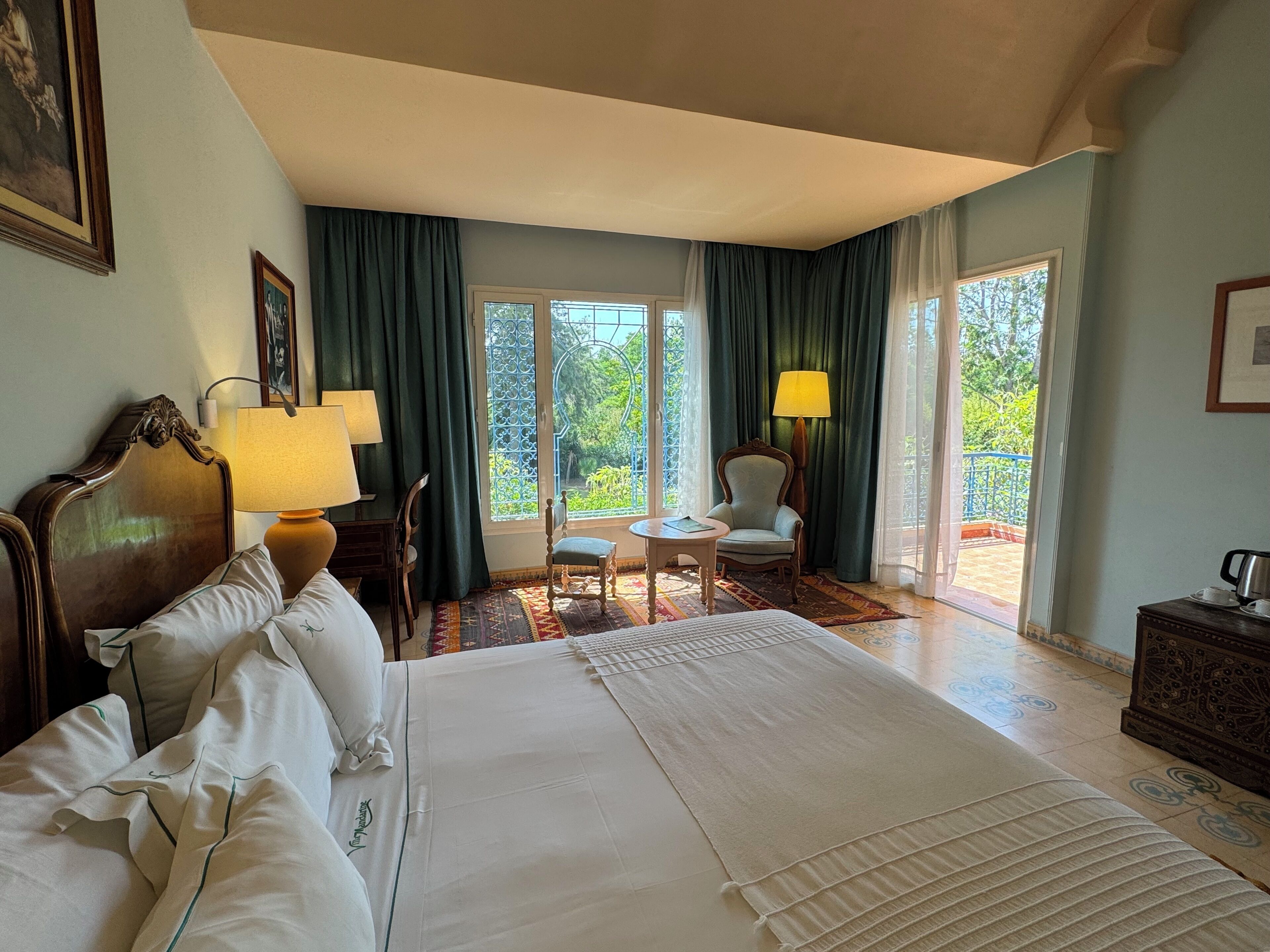 A hotel room with a large bed, two bedside lamps, a chair, a small table, and a view of greenery through the windows.