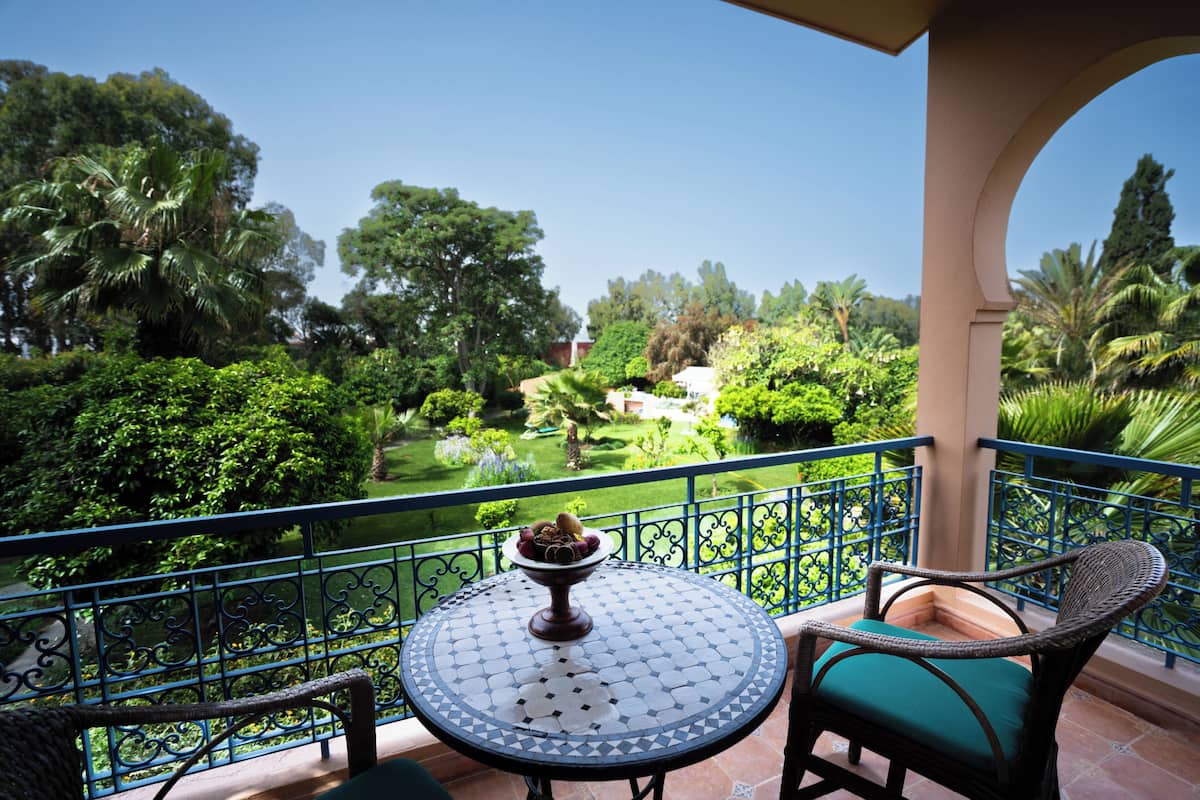 A balcony with a round table and wicker chairs, overlooking a lush garden with various trees and a fountain.