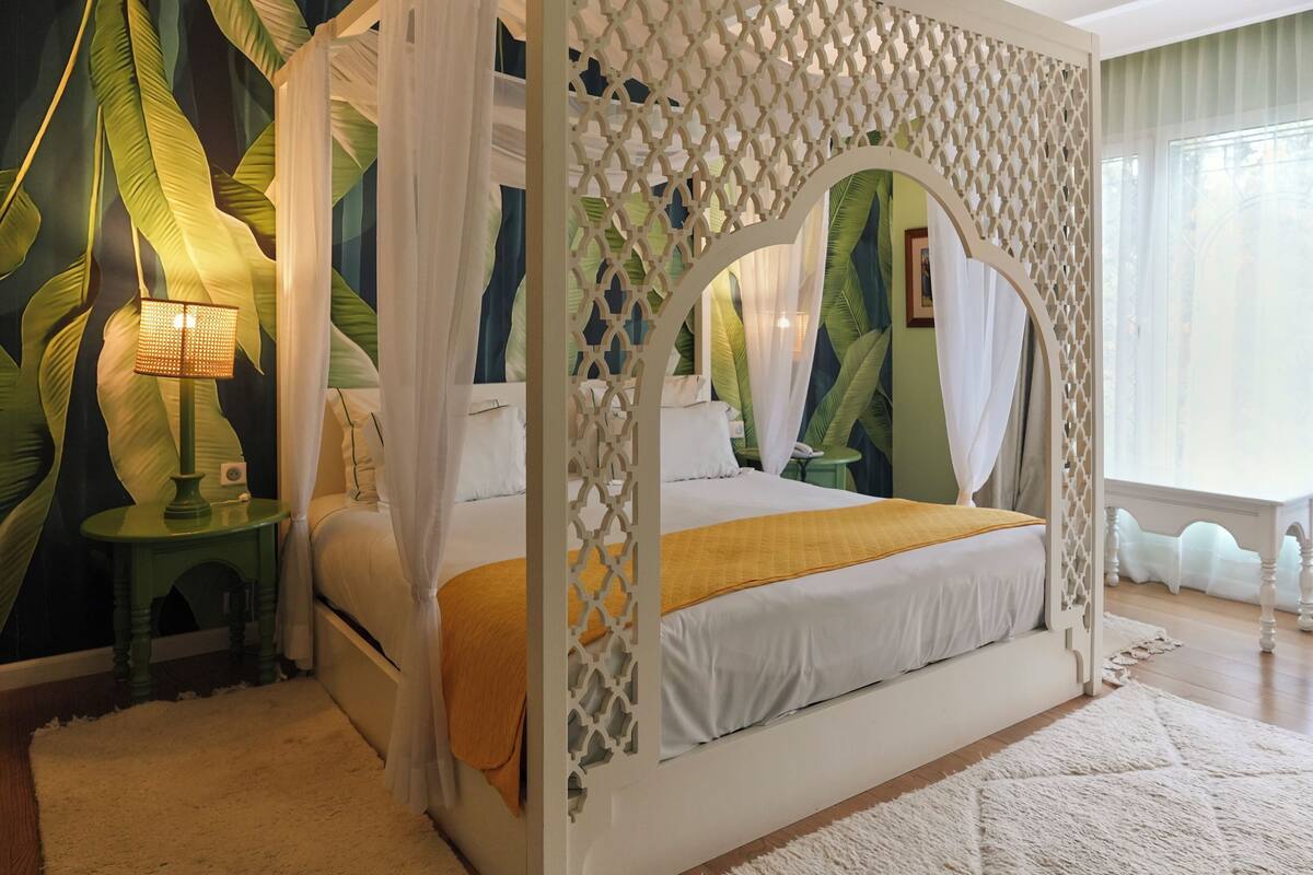 A four-poster bed with a canopy, a bedside table with a lamp, and a large window with curtains.