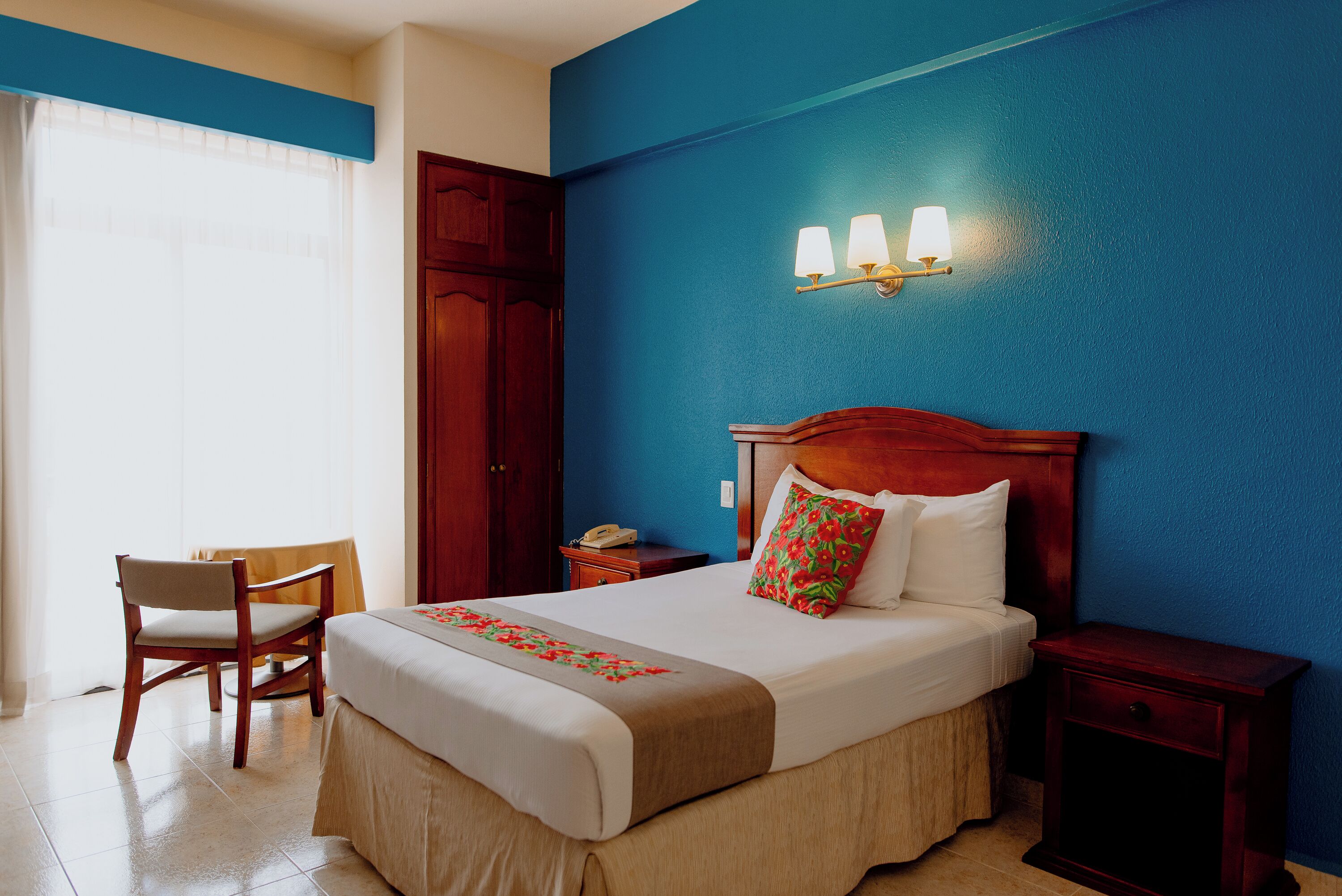 Habitación de hotel con cama, una silla, una mesita de noche y una lámpara fijada a la pared.