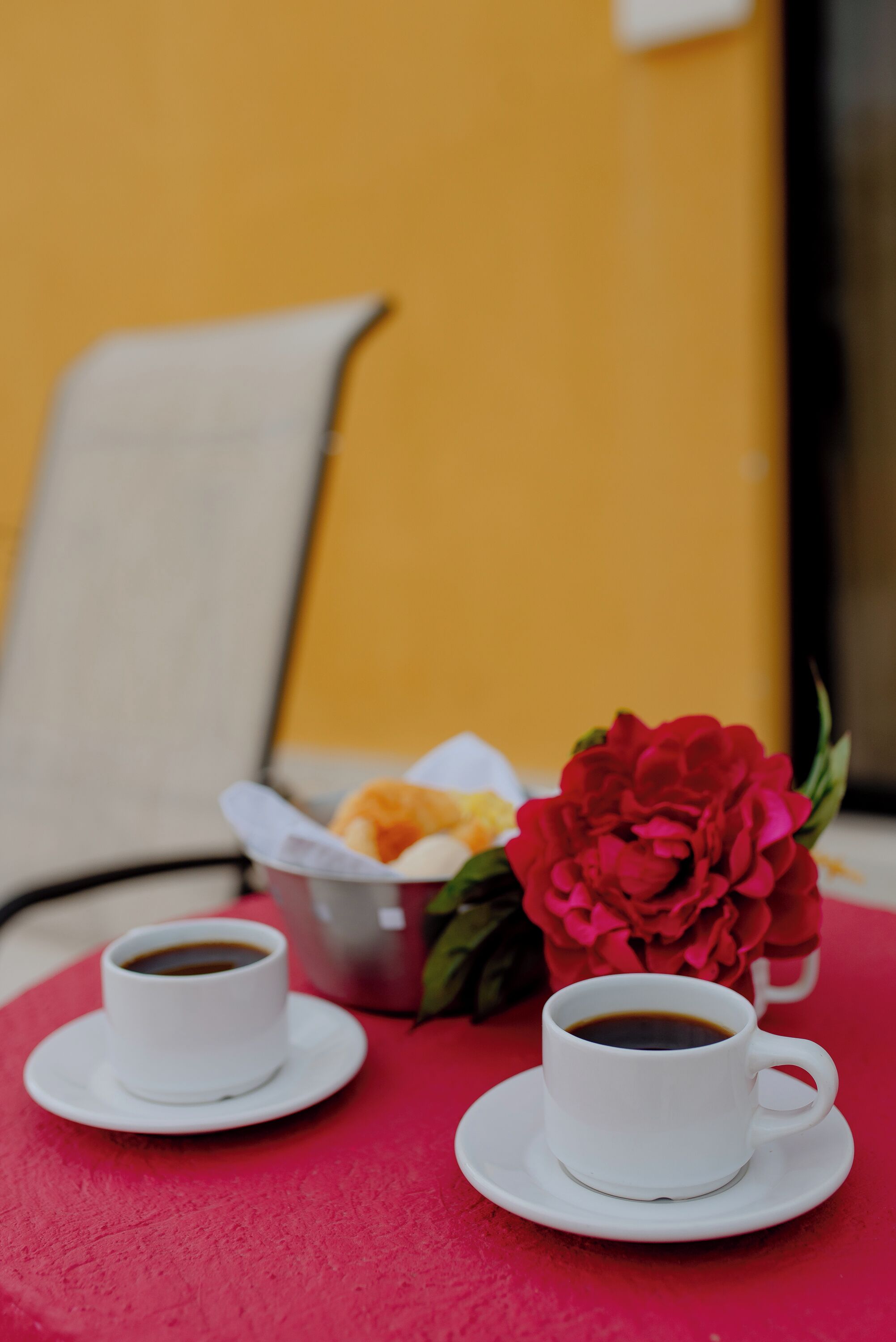 Two white cups of coffee on a red table, a basket with pastries, and a red flower.
