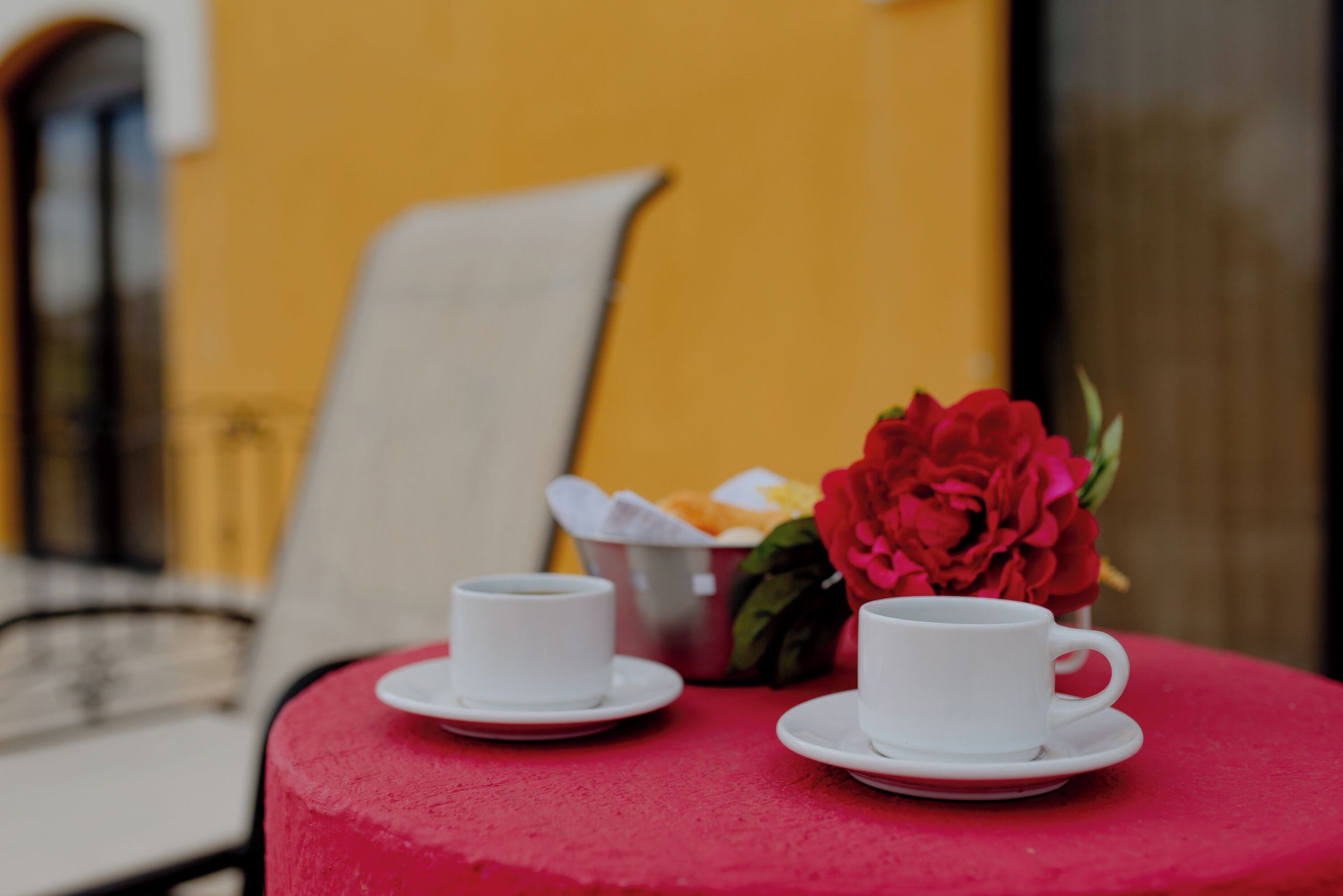 Dos tazas blancas sobre una mesa roja, con una flor roja y una cesta de frutas al fondo.