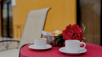 Dos tazas blancas sobre una mesa roja, con una flor roja y una cesta de frutas al fondo.