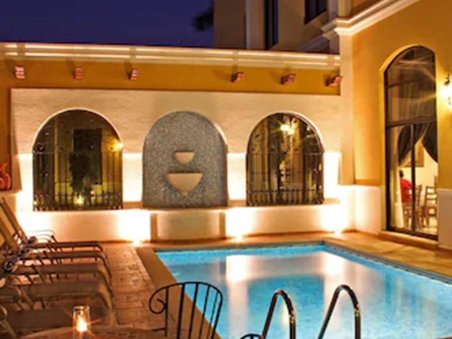Uma área de piscina com mesa e cadeiras, piso de azulejos e um edifício com janelas em arco e parede de pedra.