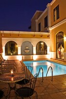 A swimming pool area with a table and chairs, a tiled floor, and a building with arched windows and a stone wall.