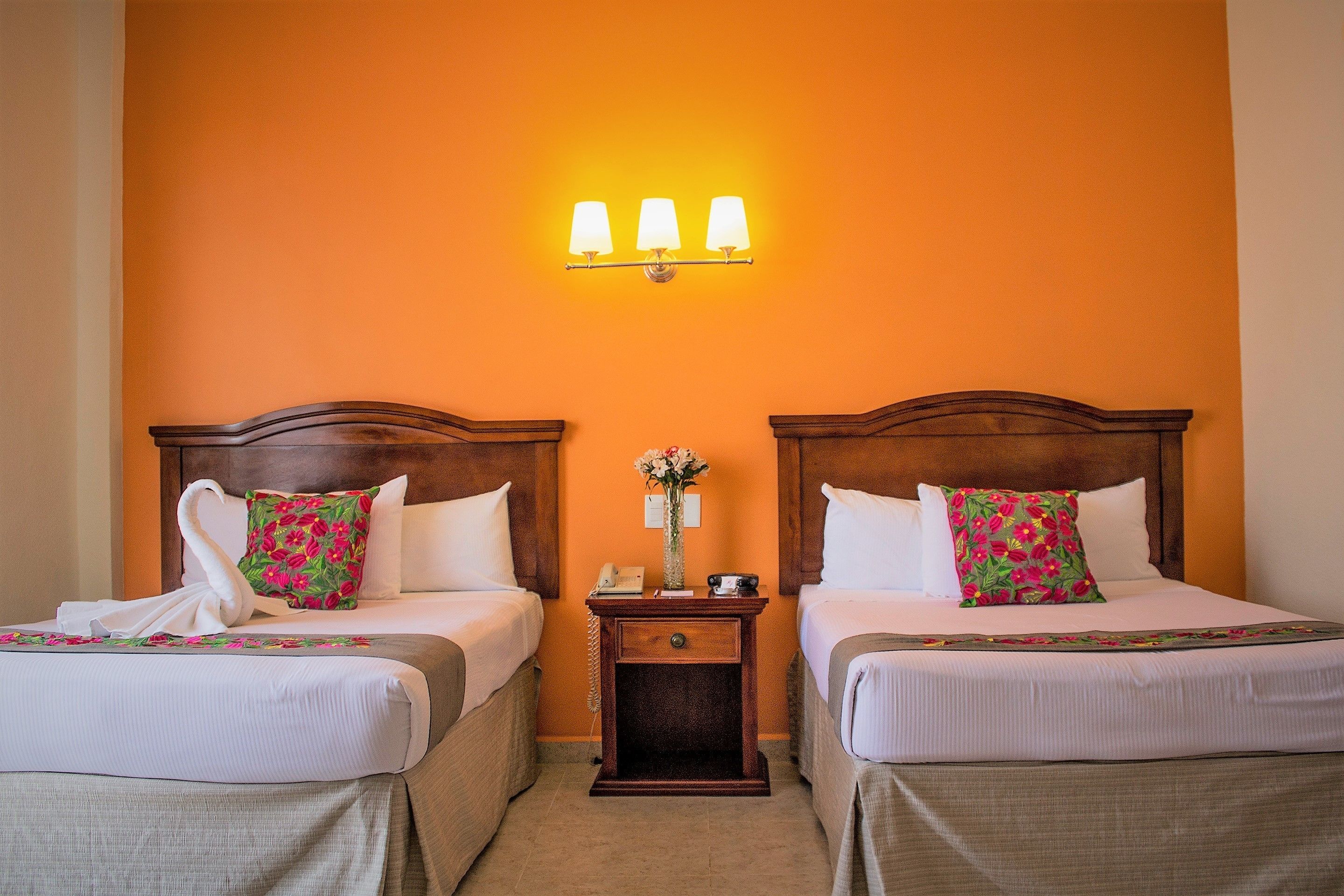 A hotel room with two beds, a nightstand with a lamp, and a wall-mounted light fixture.