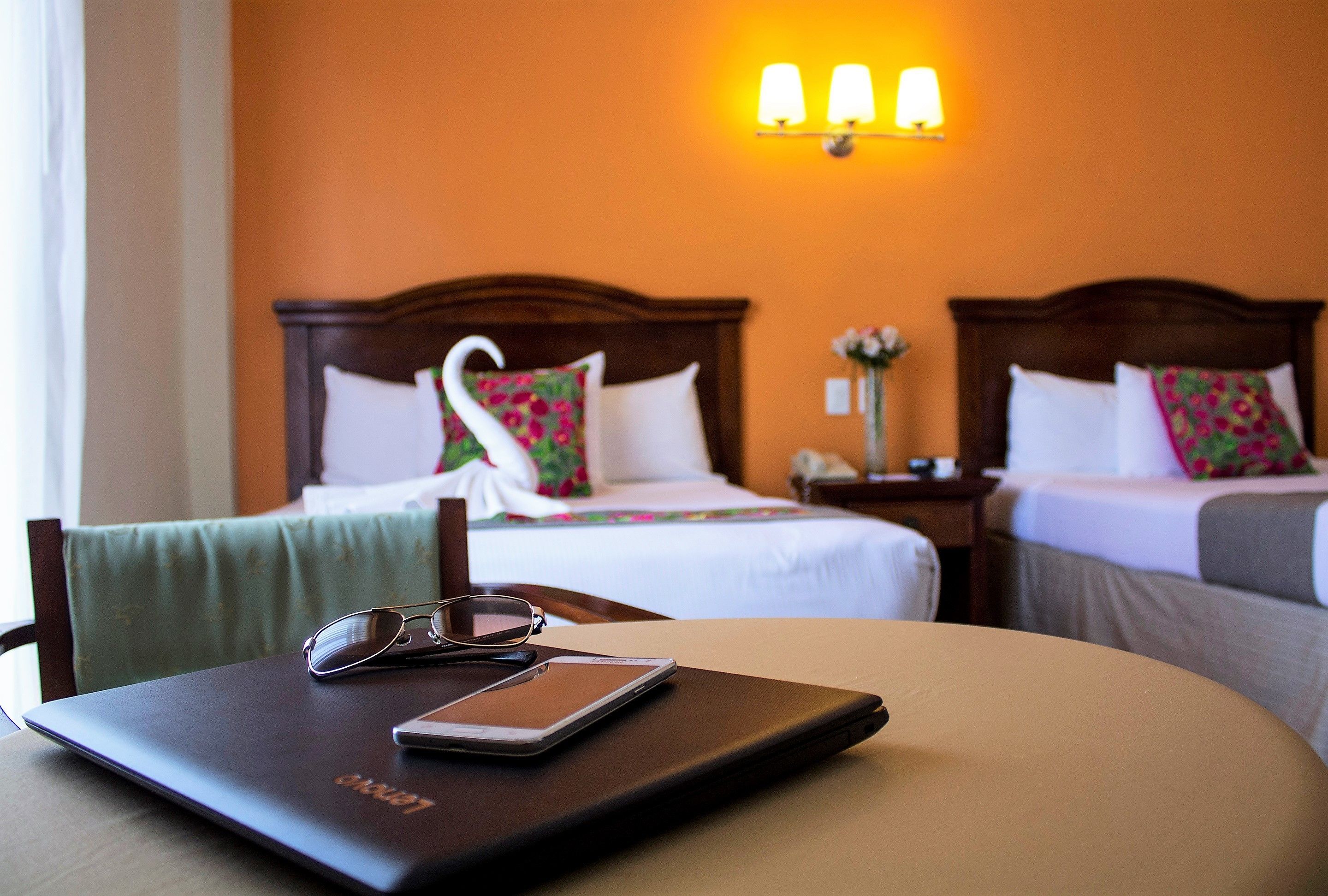 Quarto de hotel com duas camas, uma mesa com um laptop e copos, e uma luminária fixada na parede.