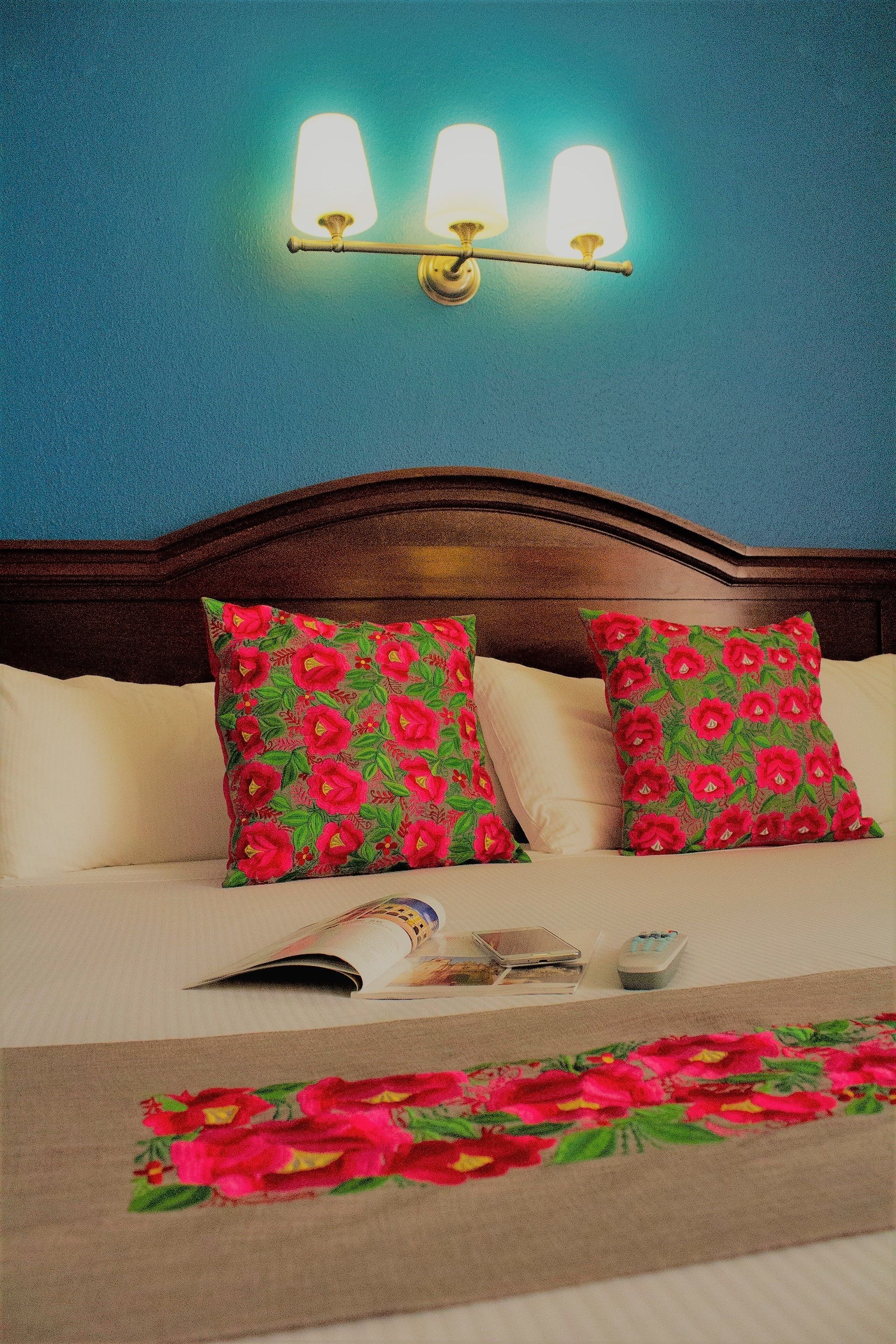 Un dormitorio con cabecera de madera, dos almohadas con estampado floral, una mesita de noche con un control remoto y una lámpara en la pared.