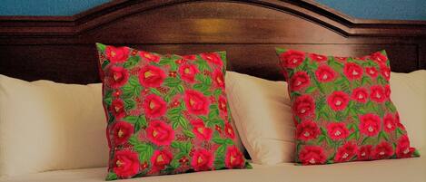 Un dormitorio con cabecera de madera, dos almohadas con estampado floral, una mesita de noche con un control remoto y una lámpara en la pared.
