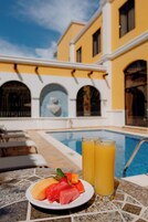 A poolside table with a plate of fruit and two glasses of orange juice, a pool, and a yellow building with arched windows in the background.