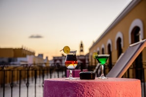 A table with two drinks and a small container, set against a sunset backdrop with buildings in the distance.