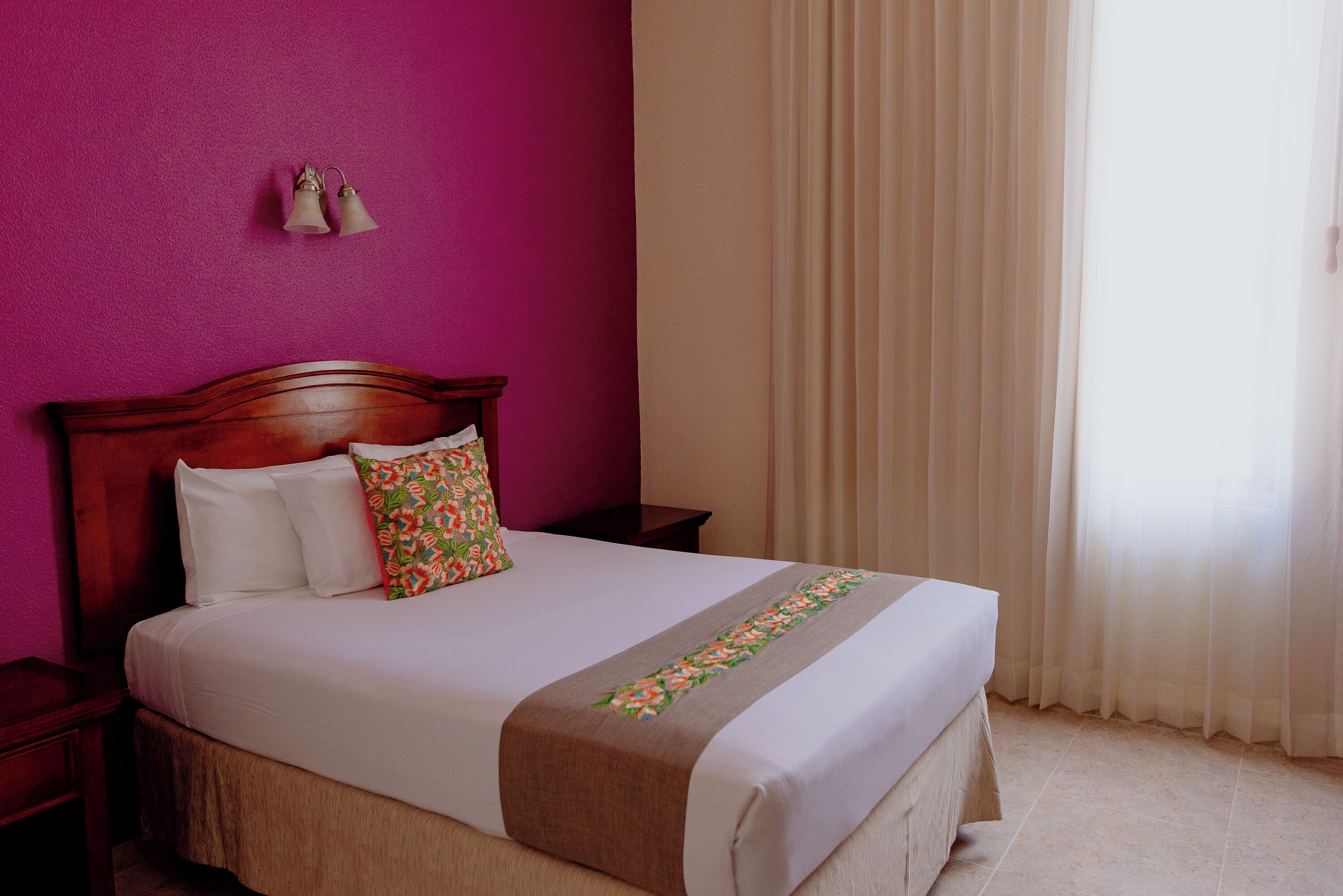 A neatly made bed with a floral pillow and a brown bedspread, a wooden nightstand with a lamp, and a window with sheer curtains.