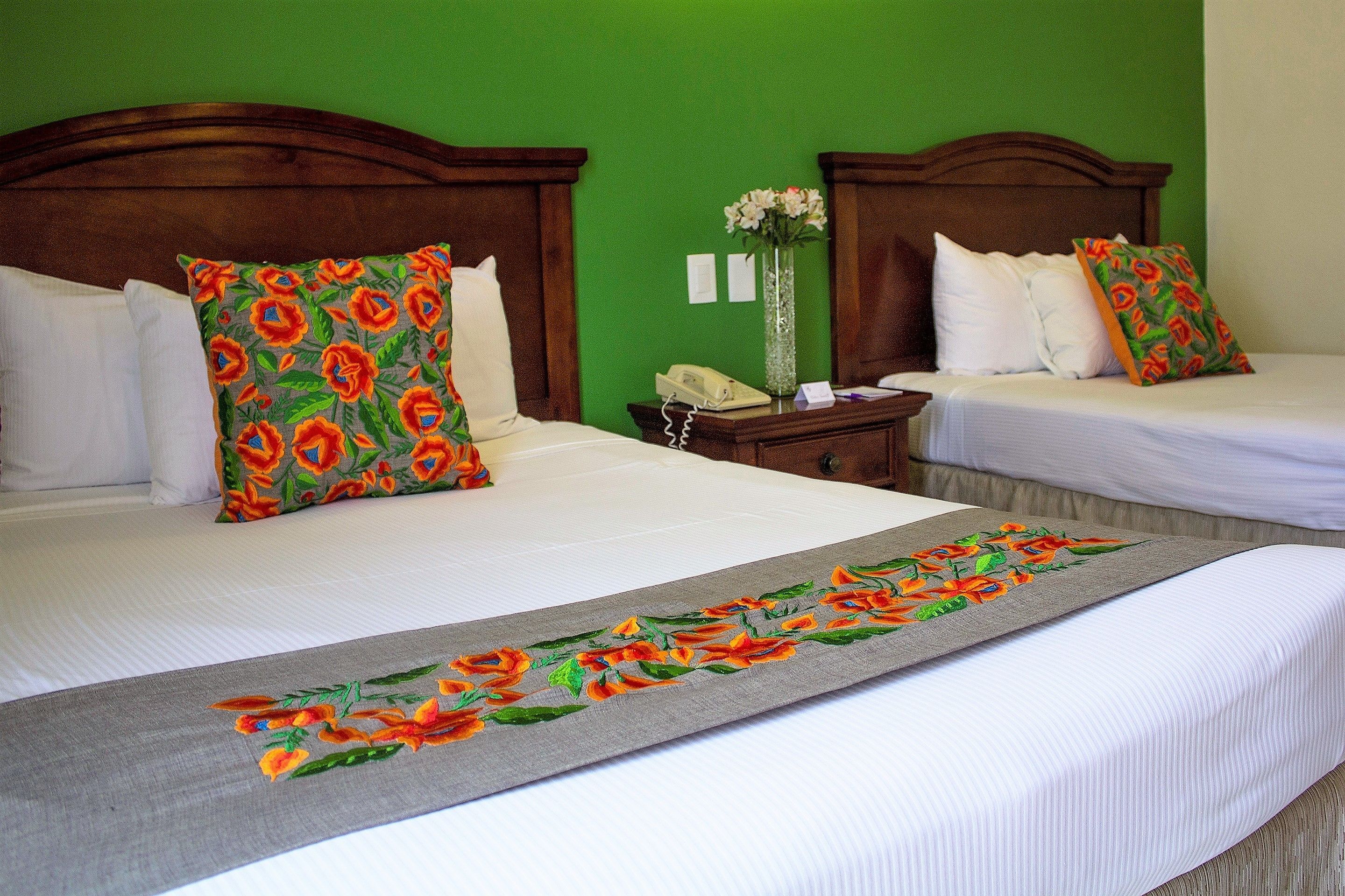 A hotel room with two beds, each with white bedding and floral-patterned pillows. There is a wooden headboard, a nightstand with a telephone, and a vase with flowers on the wall.