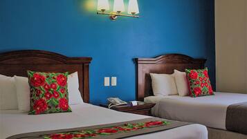 A hotel room with two beds, each with a floral pillow, a bedside table with a lamp, and a wall-mounted light fixture.
