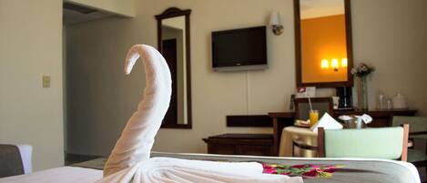 A hotel room with a neatly folded towel on the bed, a television mounted on the wall, a mirror, and a desk with a chair.
