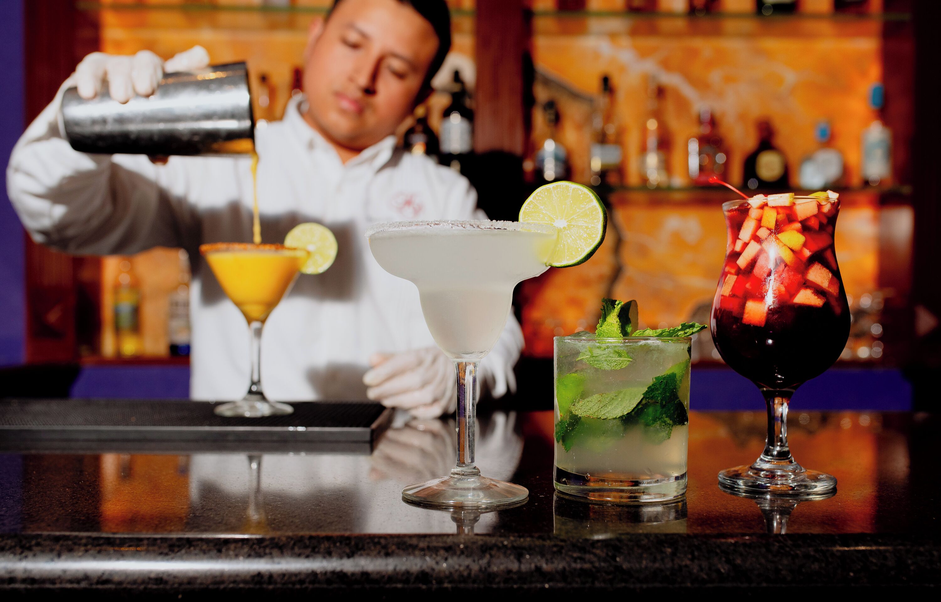 A bartender pouring a drink into a margarita glass, with other cocktails on the bar.