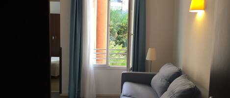 A room with a grey sofa, a window with blue curtains, a lamp, and a view of greenery outside.