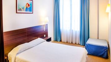 A hotel room with a wooden headboard, a bed with white linens, a blue upholstered bench, a nightstand with a lamp, and a painting on the wall.