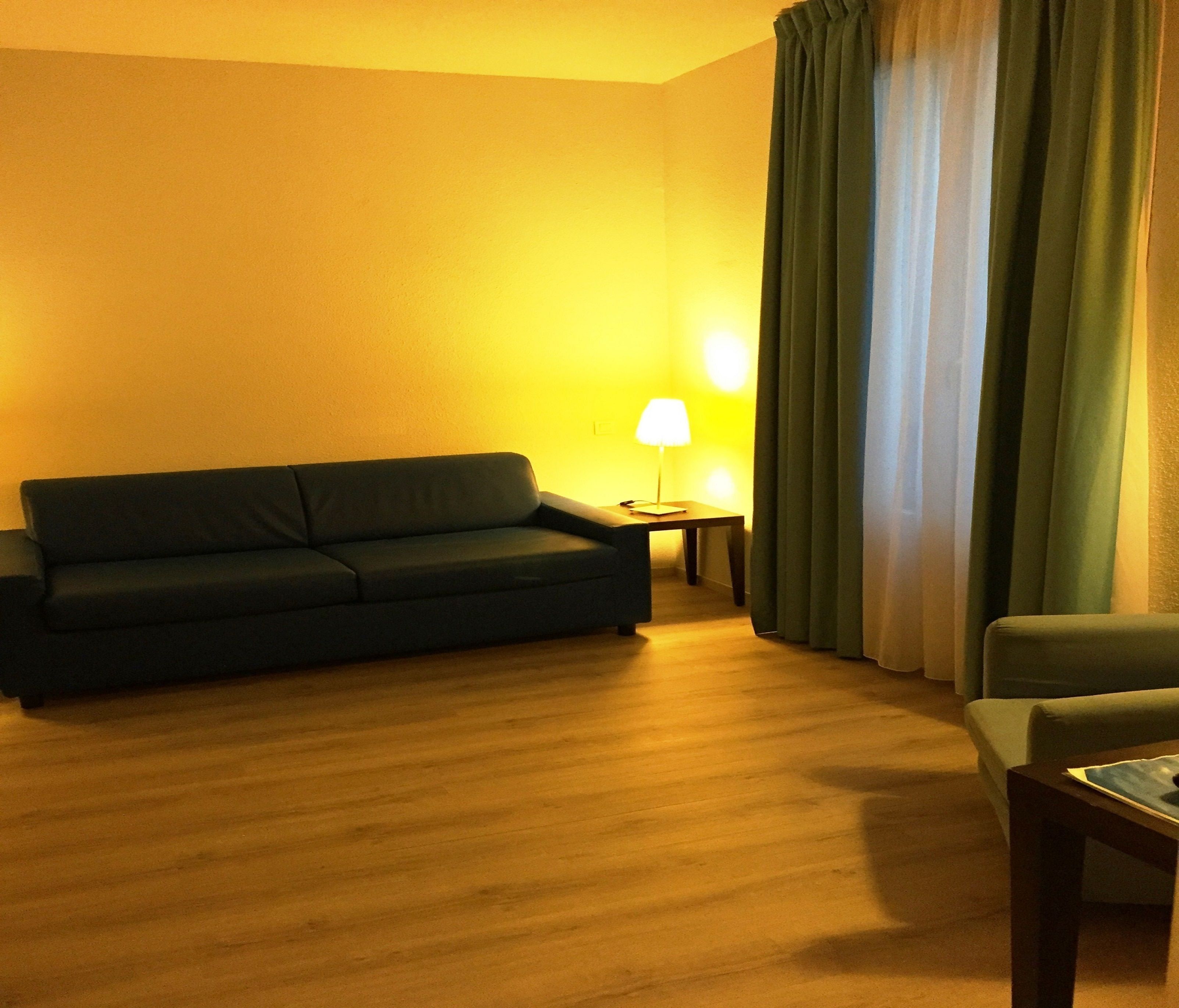 A room with a dark green sofa, a small table with a lamp, and a chair.