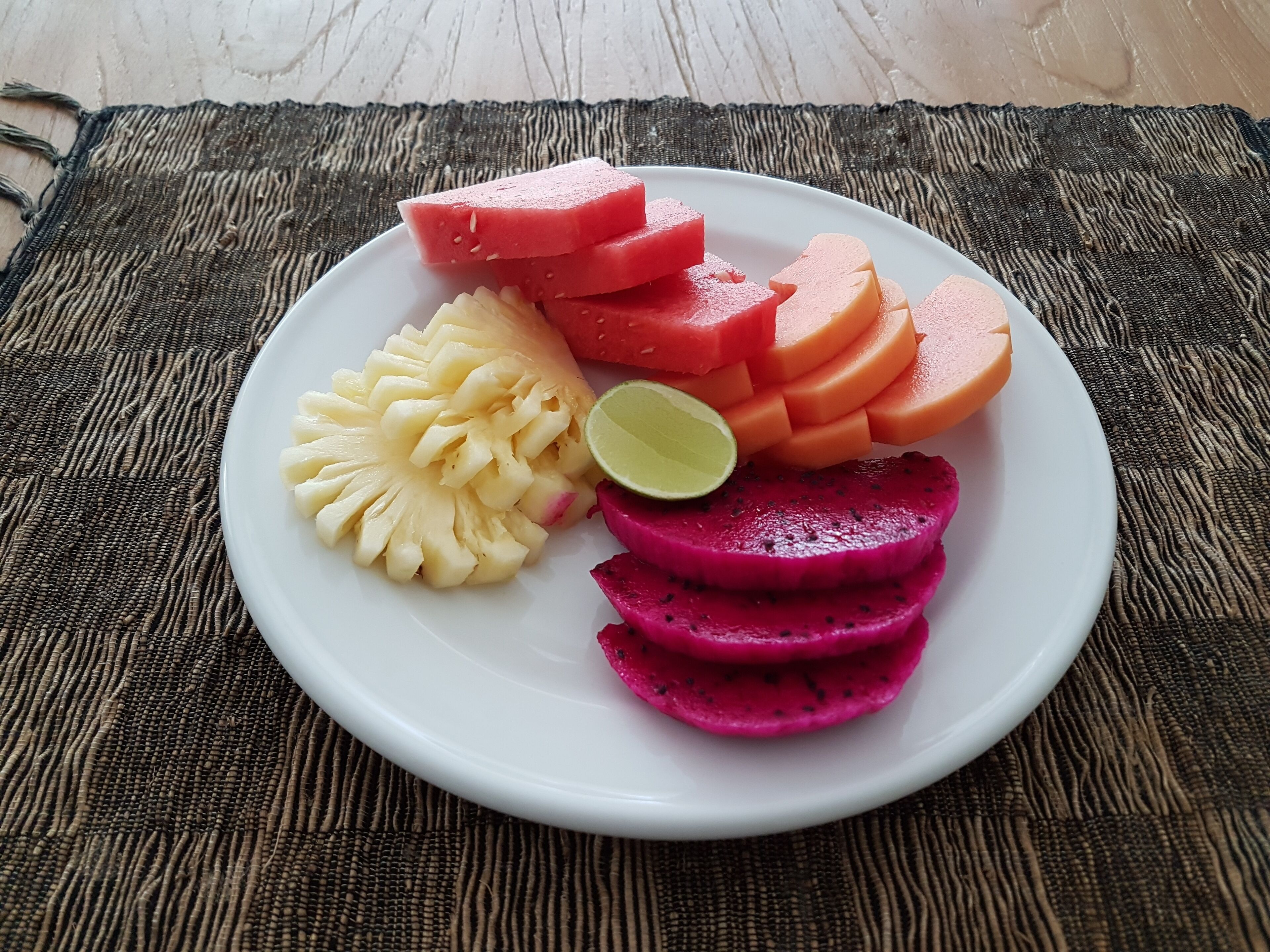 Una fuente con sandía, piña, pitahaya y lima sobre una estera tejida.