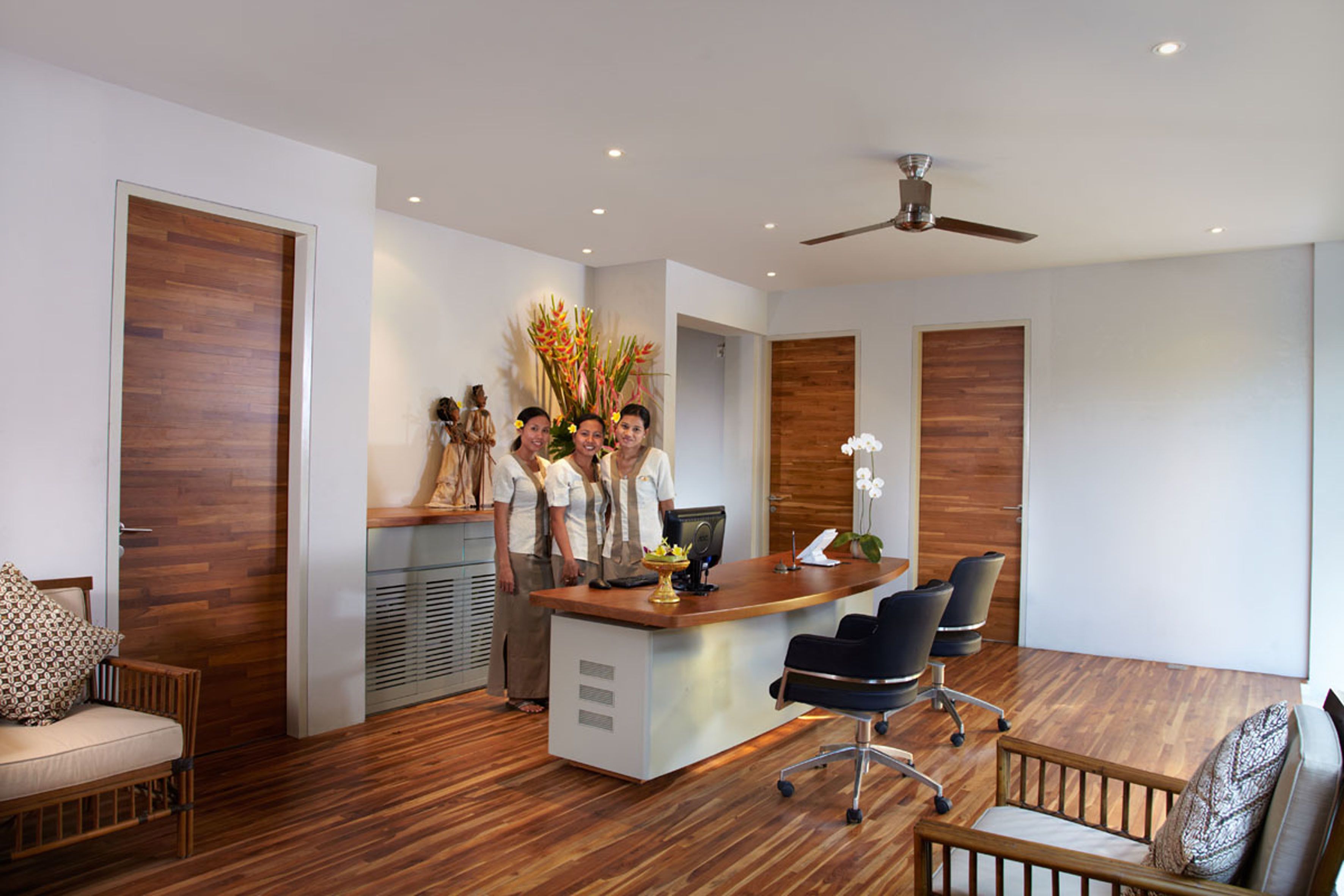 A modern office with a reception desk, wooden flooring, a ceiling fan, and a seating area with a sofa and chairs.