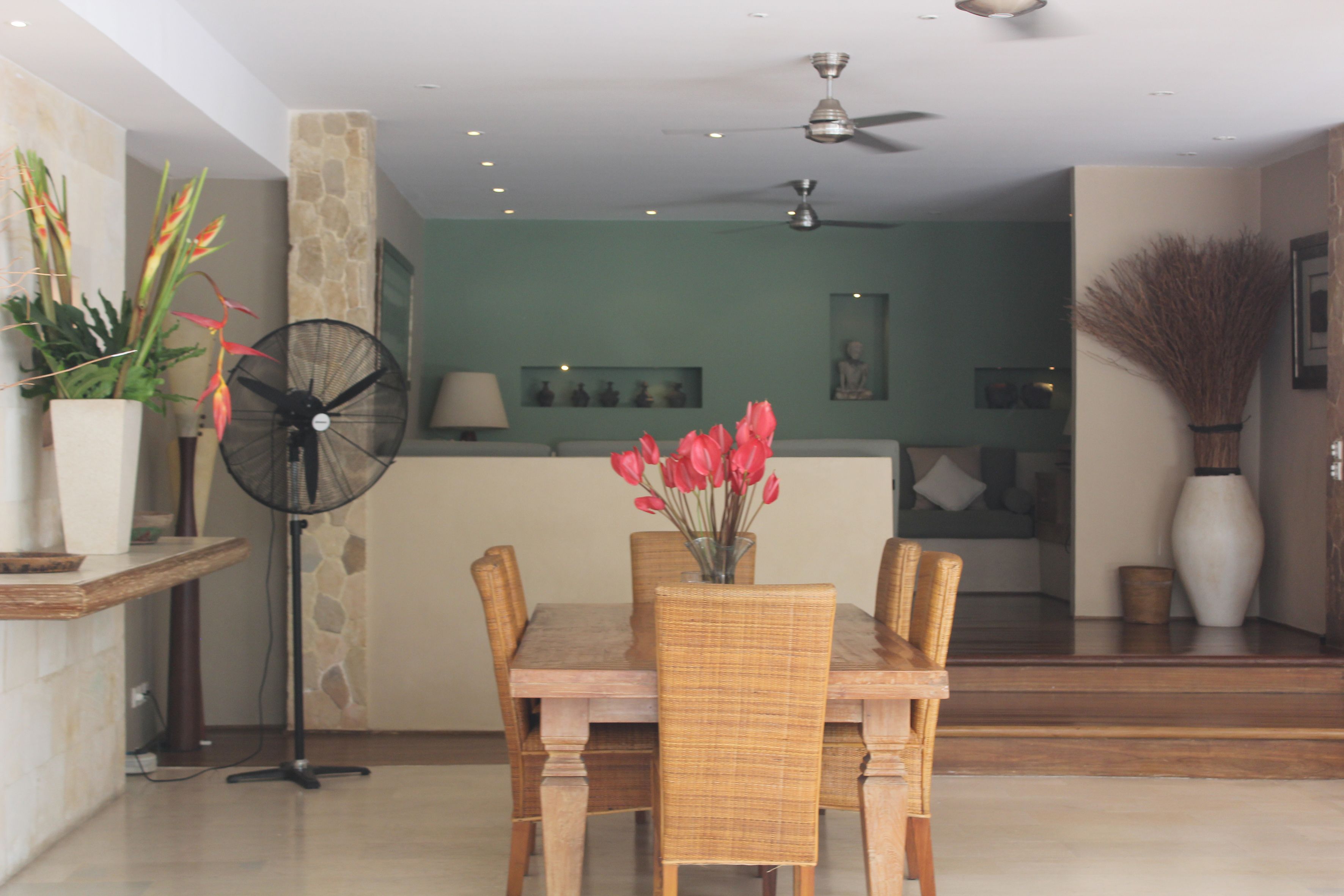 A dining area with a wooden table and chairs, a fan, and a vase with red flowers.