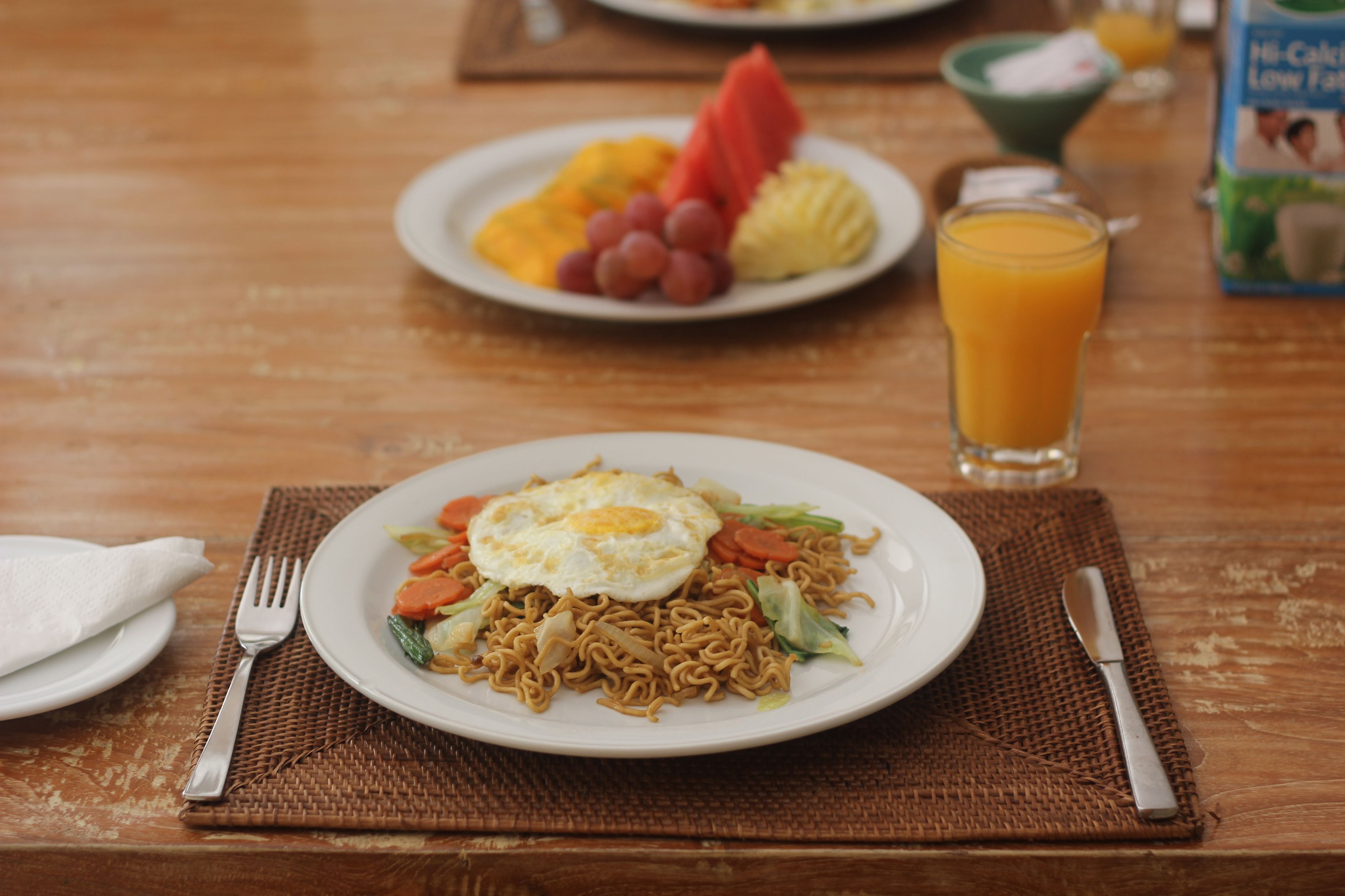 A plate of noodles with vegetables and an egg, a glass of orange juice, and a bowl of fruit on a wooden table.