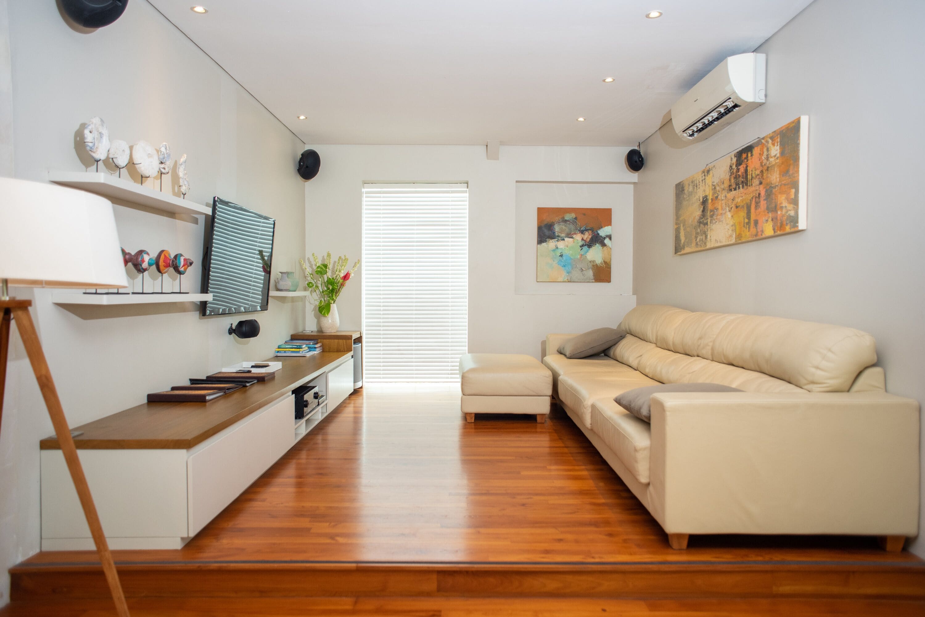 A modern living room with a sofa, a coffee table, and a TV. There is a window with blinds, a painting on the wall, and a plant on the side table.