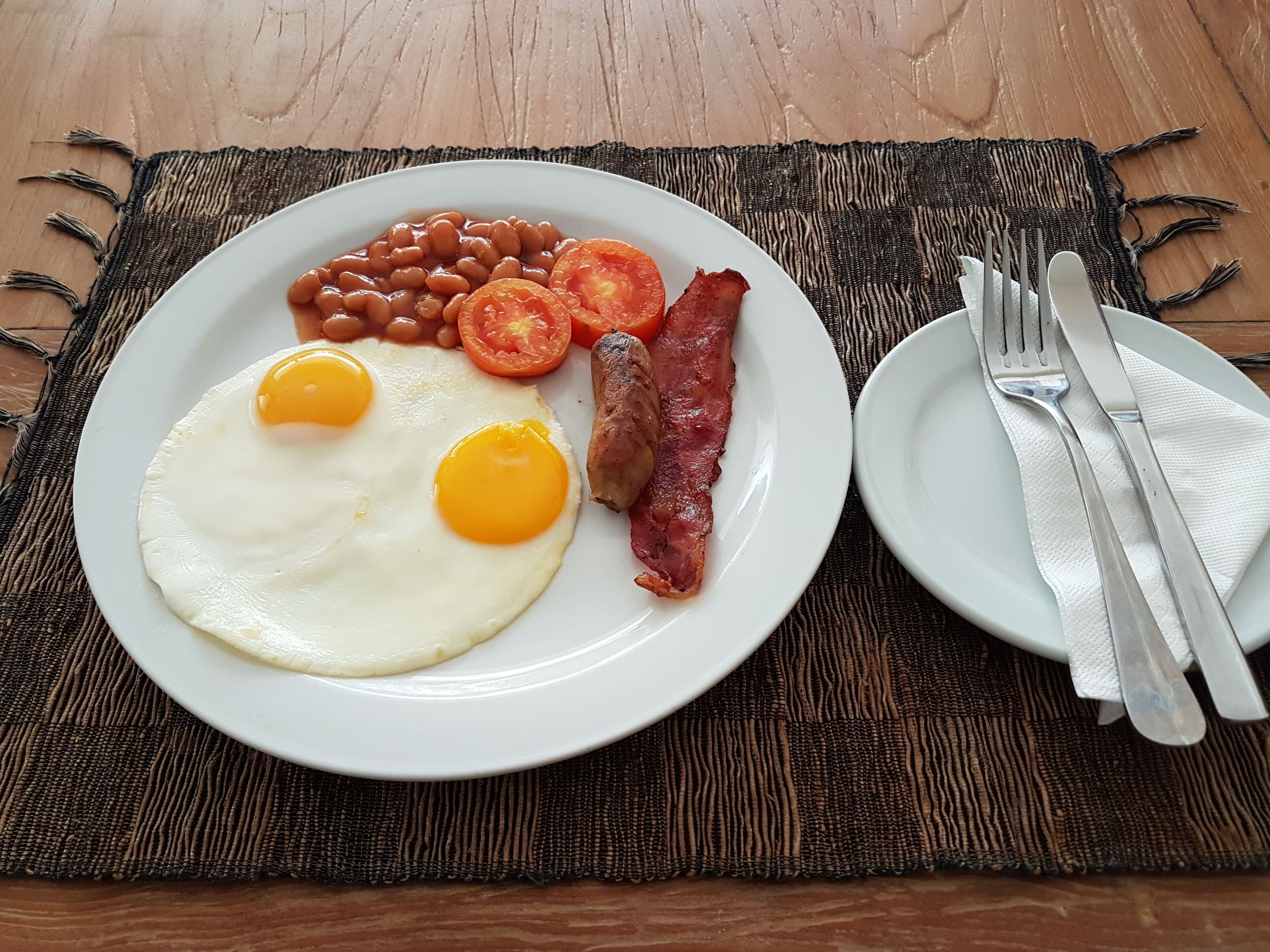 Ein Teller mit Eiern, Speck, Tomaten und Bohnen auf einer gewebten Tischmatte.