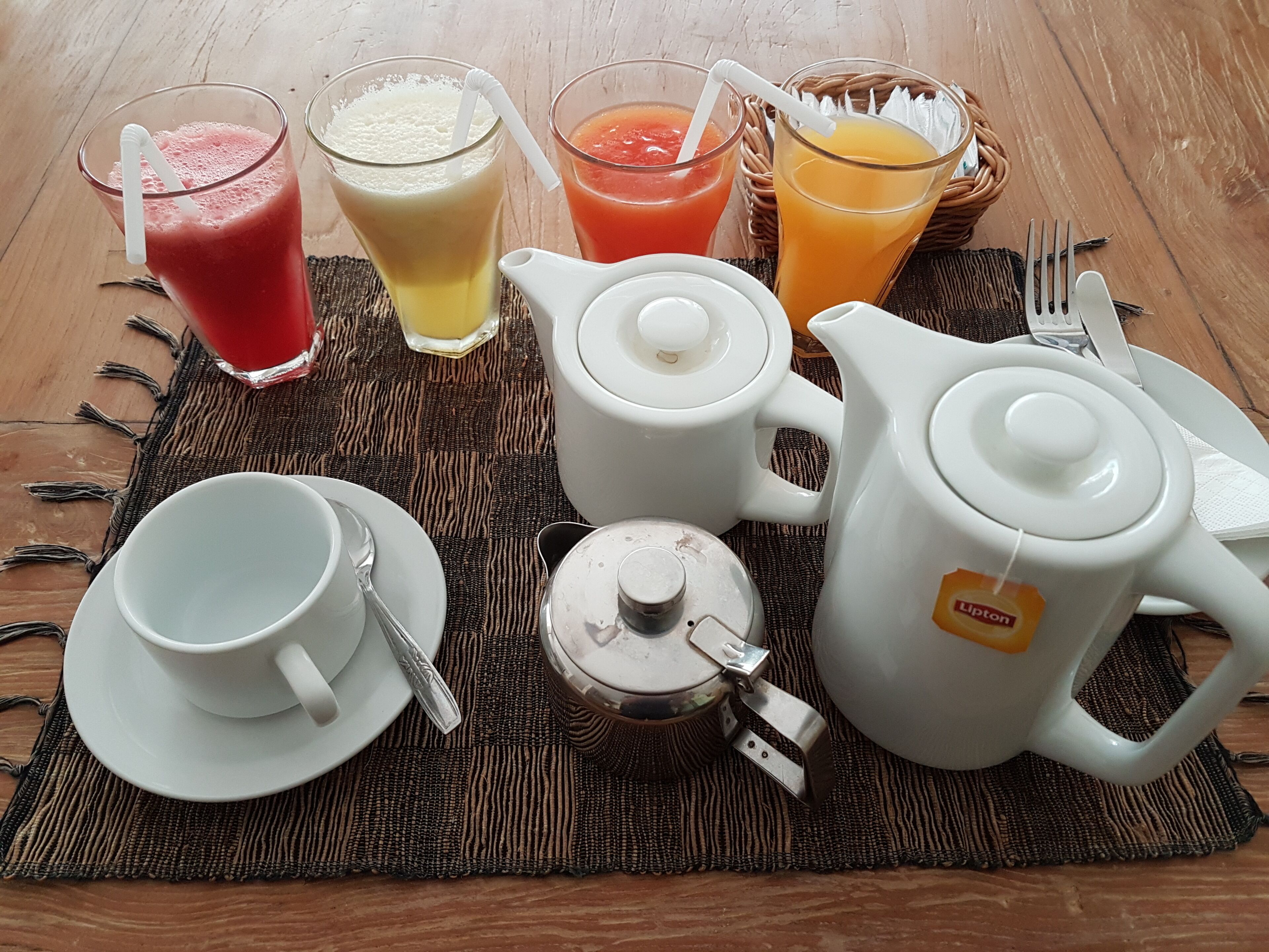 Ein Frühstücksarrangement mit Teekanne, Tassen, Krug und verschiedenen Getränken auf einer gewebten Unterlage.