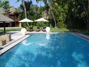 A swimming pool with lounge chairs and umbrellas, surrounded by palm trees and a building with a thatched roof. - Sienna Villas Seminyak (Seminyak)