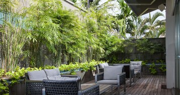 Uma área de estar externa com mobiliário de vime e um exuberante fundo verde de bambu e plantas tropicais.
