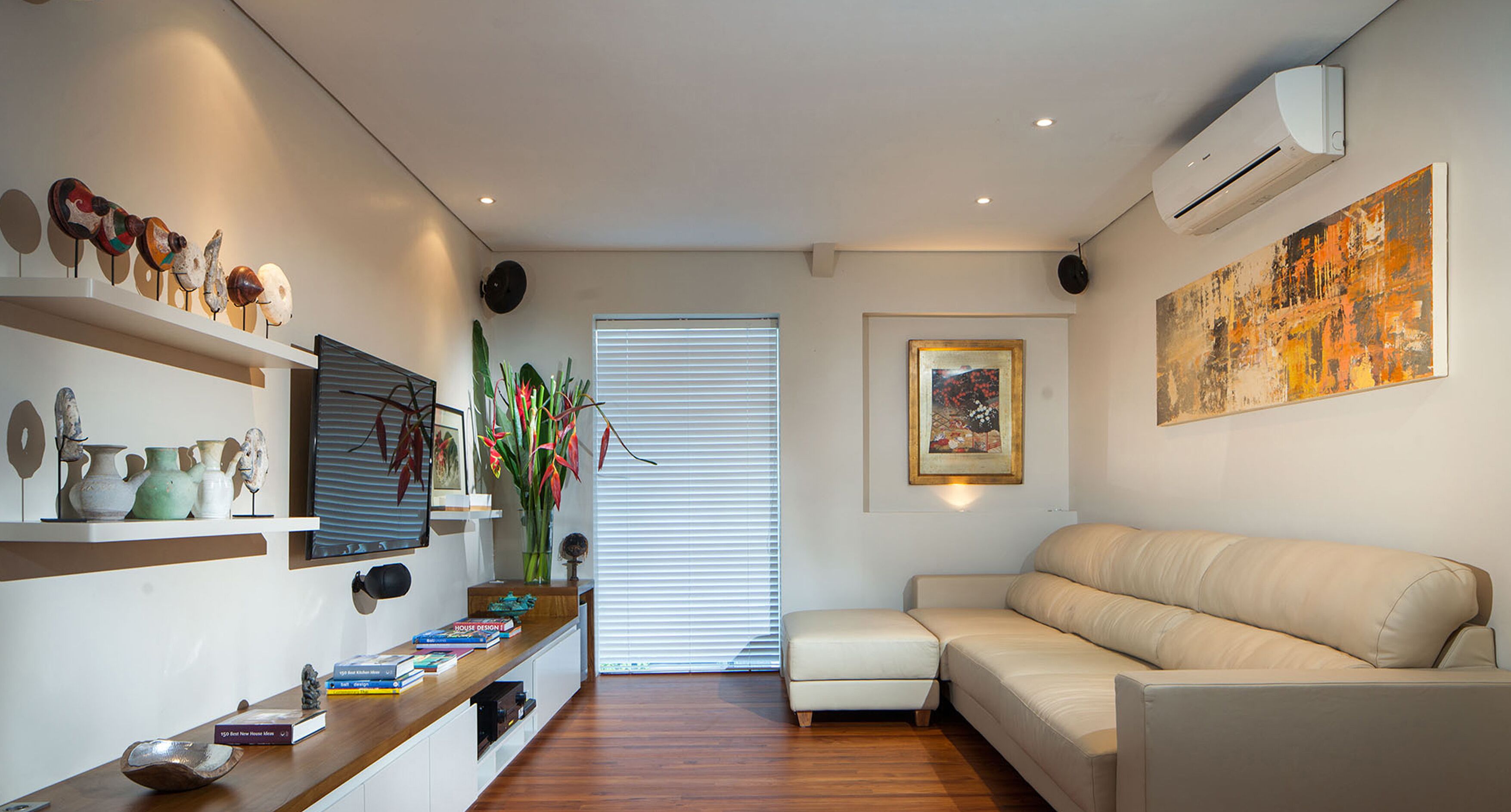 Una sala de estar moderna con un sofá, una mesa de centro y elementos decorativos. Se observa una ventana con persianas, un aire acondicionado de pared y un cuadro.