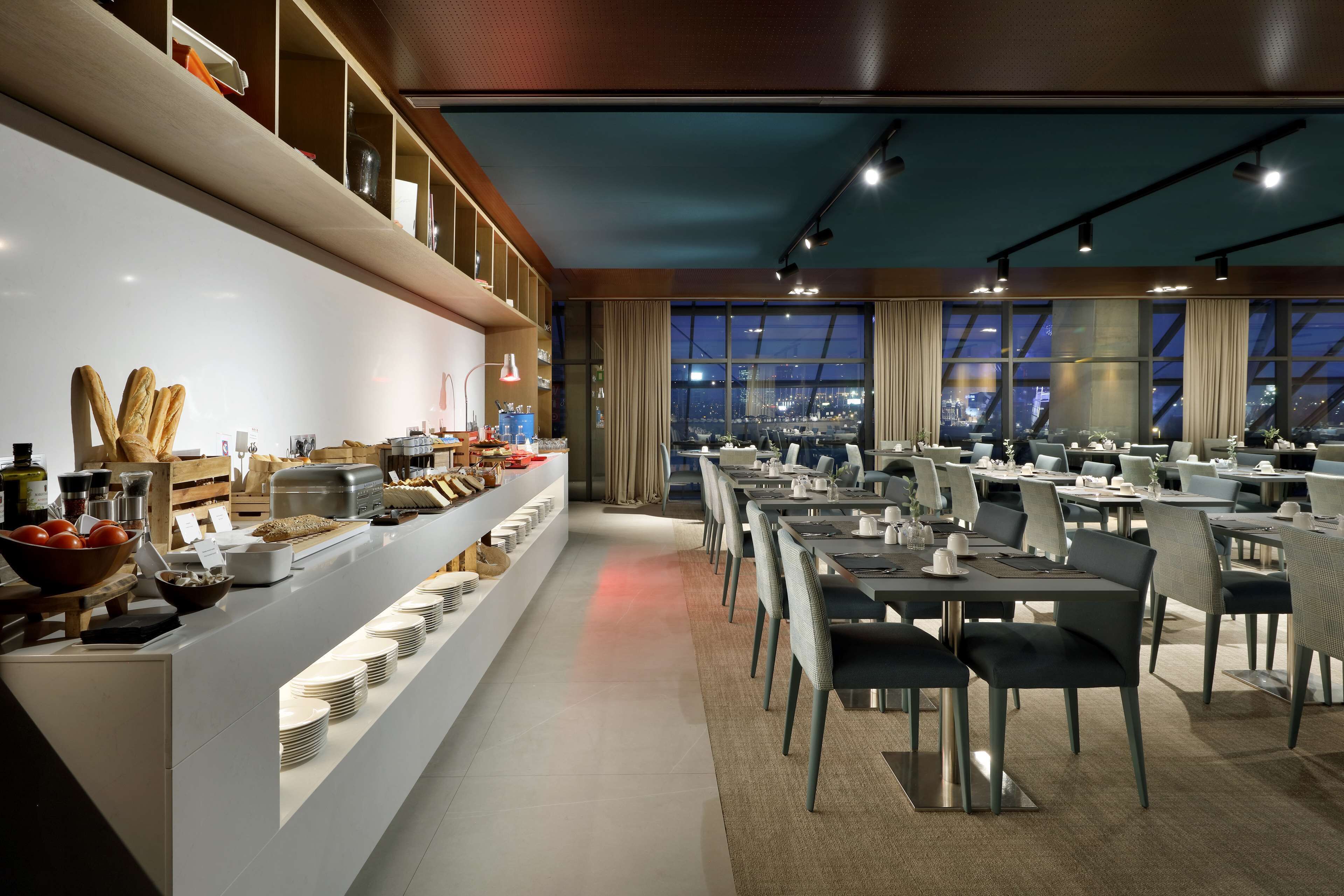 Uma área de refeições moderna com um buffet à esquerda, amplas janelas com vista para a cidade e mesas e cadeiras bem organizadas.