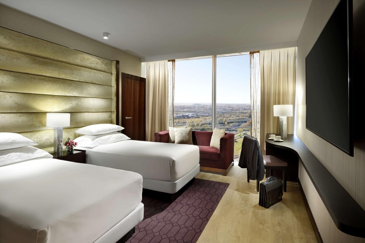 Ein Hotelzimmer mit zwei Betten, einem Schreibtisch, einem Fernseher und einem großen Fenster mit Blick auf die Stadt.