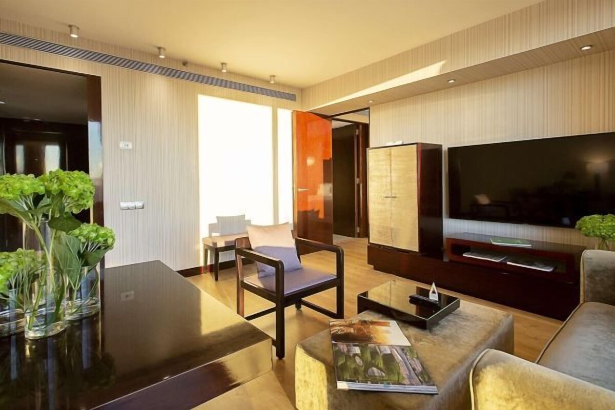 A modern living room with a flat-screen TV, a sofa, a coffee table, and a dining table. There are potted plants and a bookshelf.