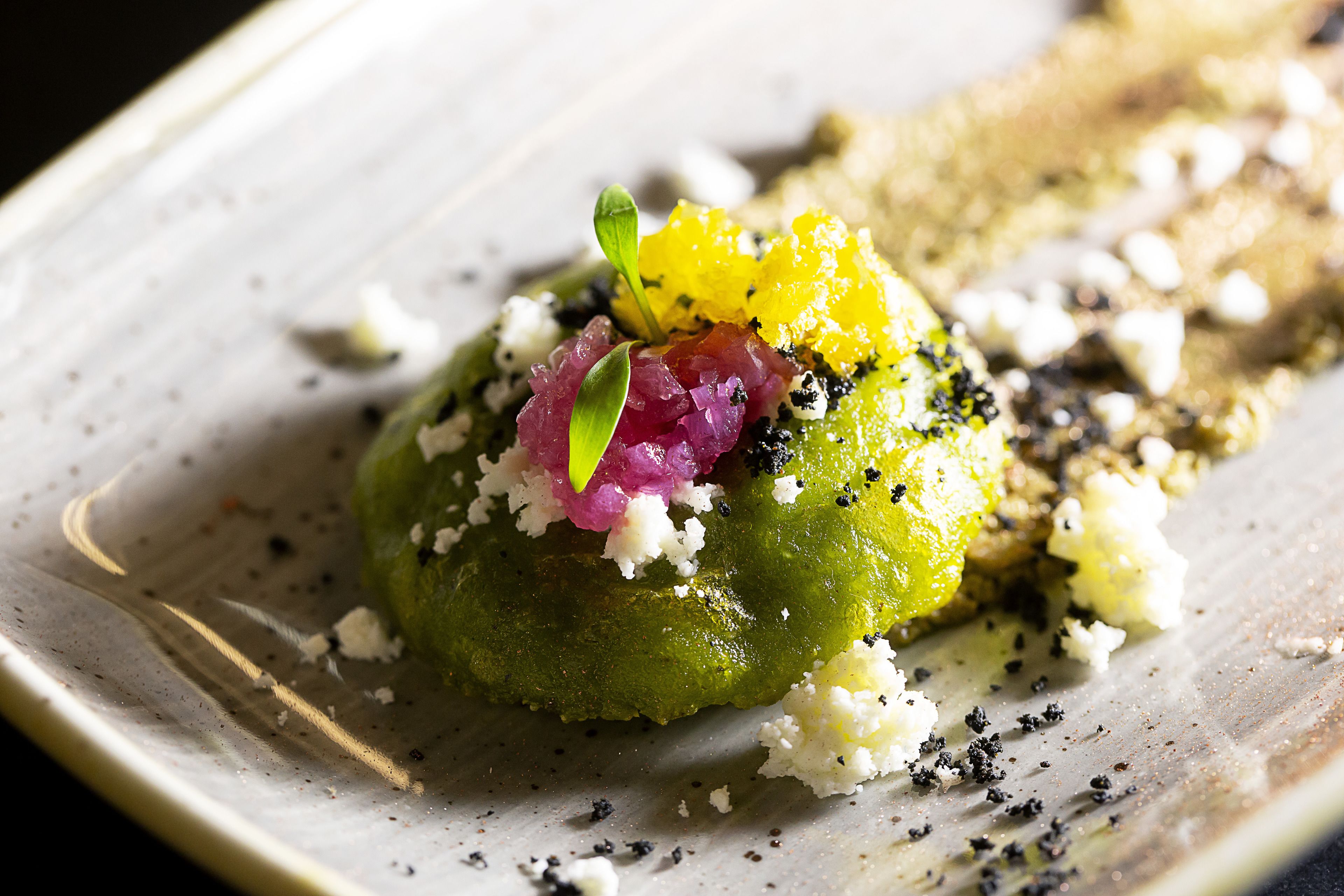 Un plato con un alimento verde, posiblemente verdura o fruta, cubierto con ingredientes rojos y amarillos, y queso blanco desmenuzado.