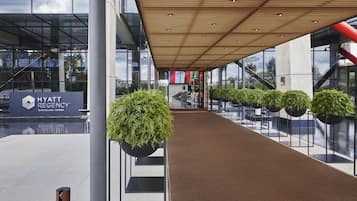 Entrada moderna de hotel com fachada de vidro, placa indicando Hyatt Regency Barcelona Tower e galeria coberta com vasos de plantas.