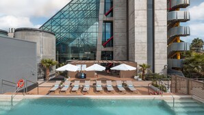 Un bâtiment moderne doté d’une façade en verre, un espace piscine avec des chaises longues et des parasols, et une vue sur la piscine.