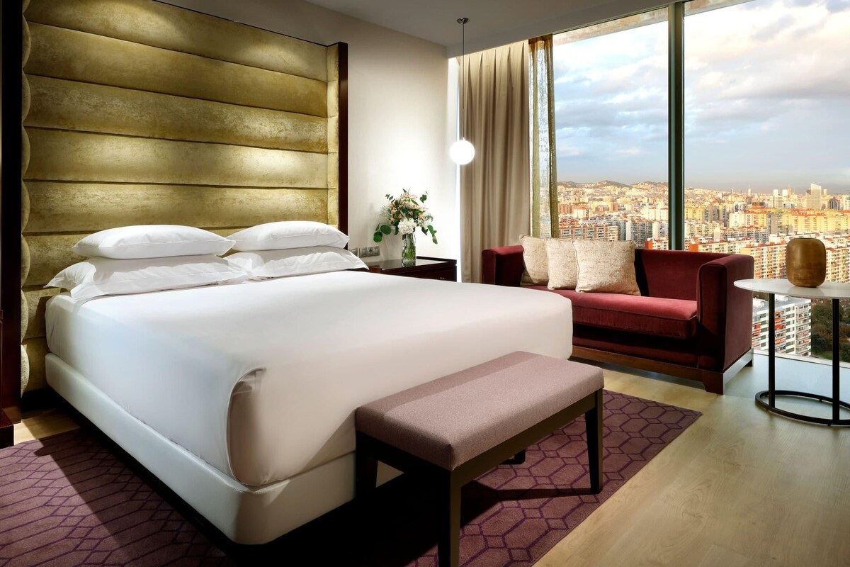 Ein modernes Hotelzimmer mit einem großen Bett, einem roten Sofa, einem kleinen Tisch und Blick auf eine Stadtlandschaft.