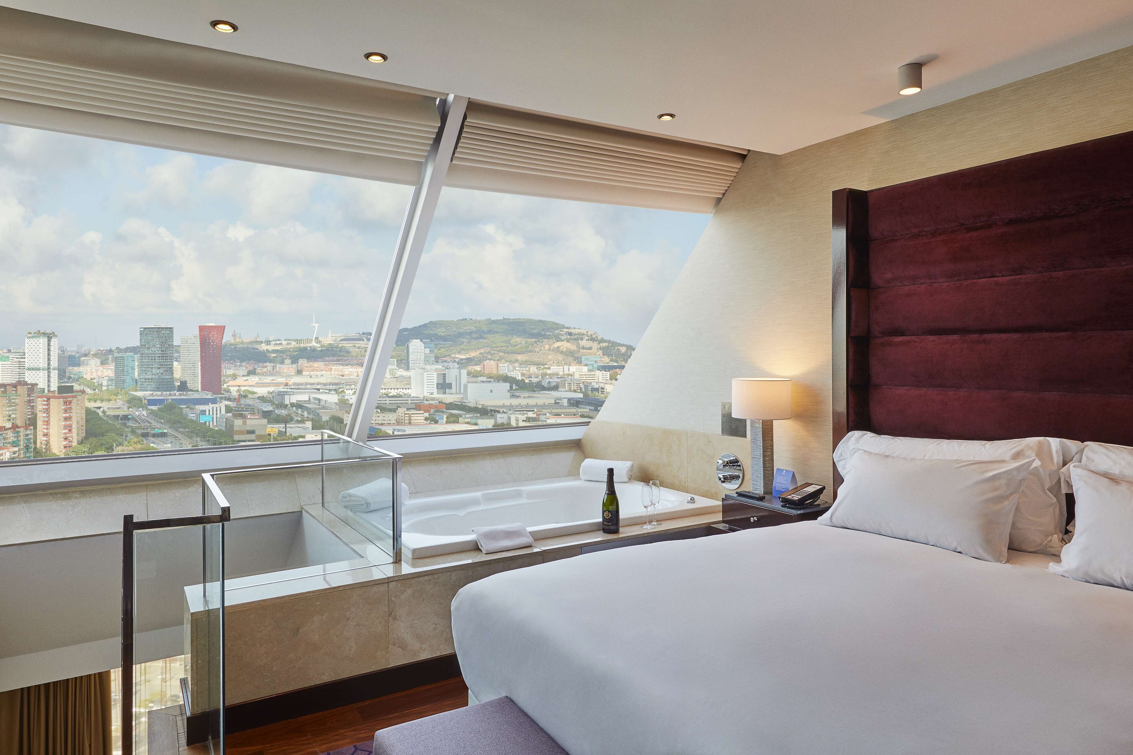 A hotel room with a large bed, a glass balcony, a city view, and a red headboard.