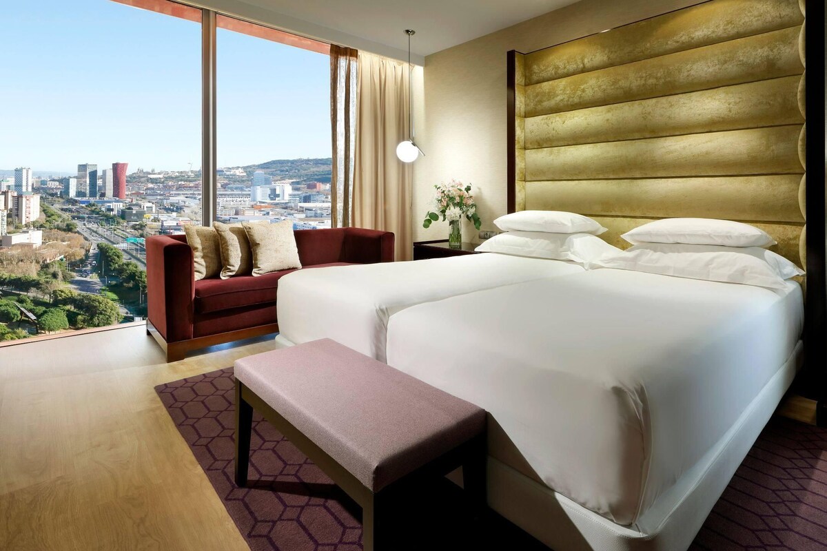 Ein Hotelzimmer mit einem großen Bett, einer Couch, einer Bank, einem Nachttisch und Blick auf die Stadt.